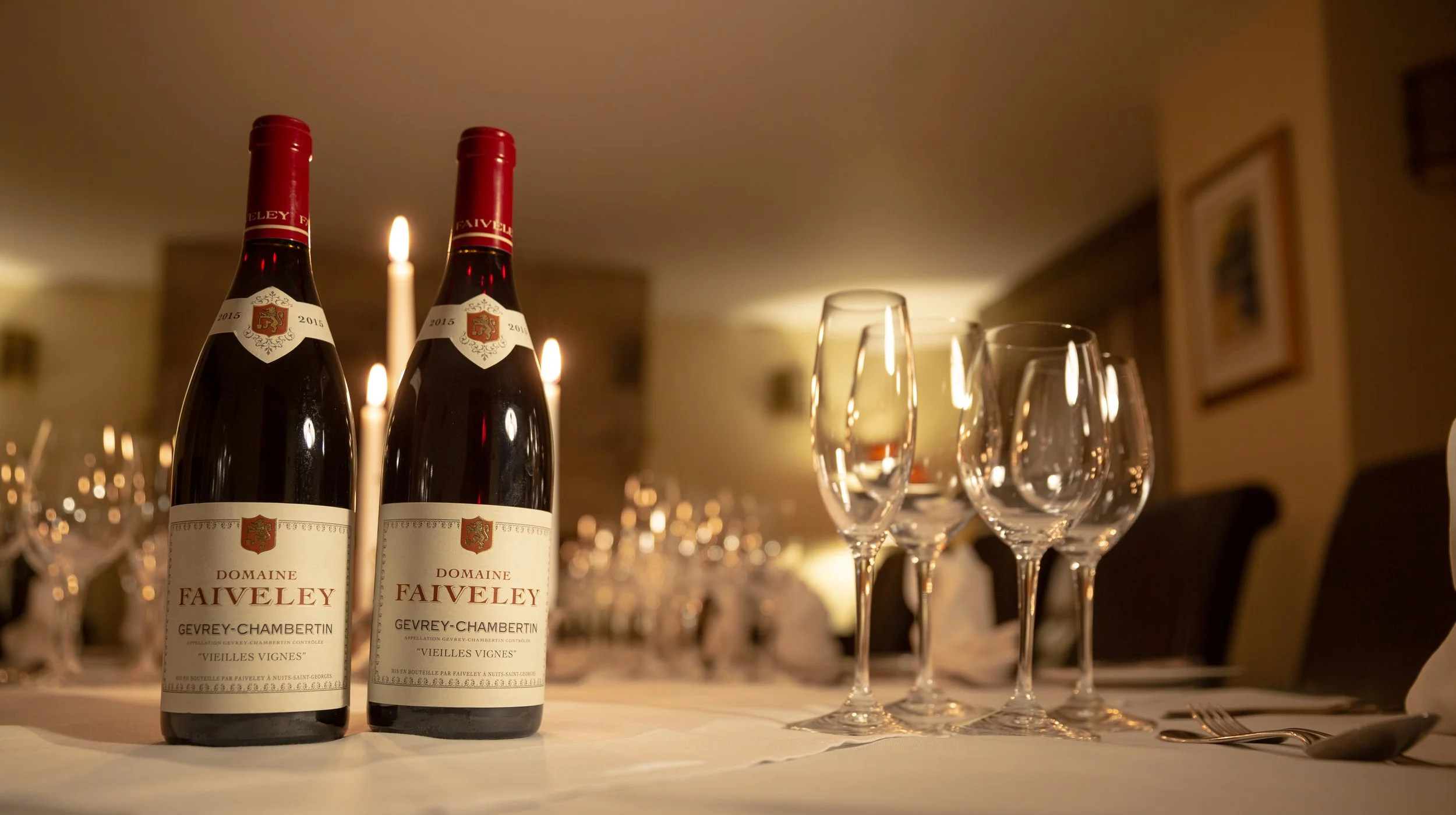 Two bottles of Domaine Faiveley red wine next to empty champagne glasses on a table in a dimly lit room.