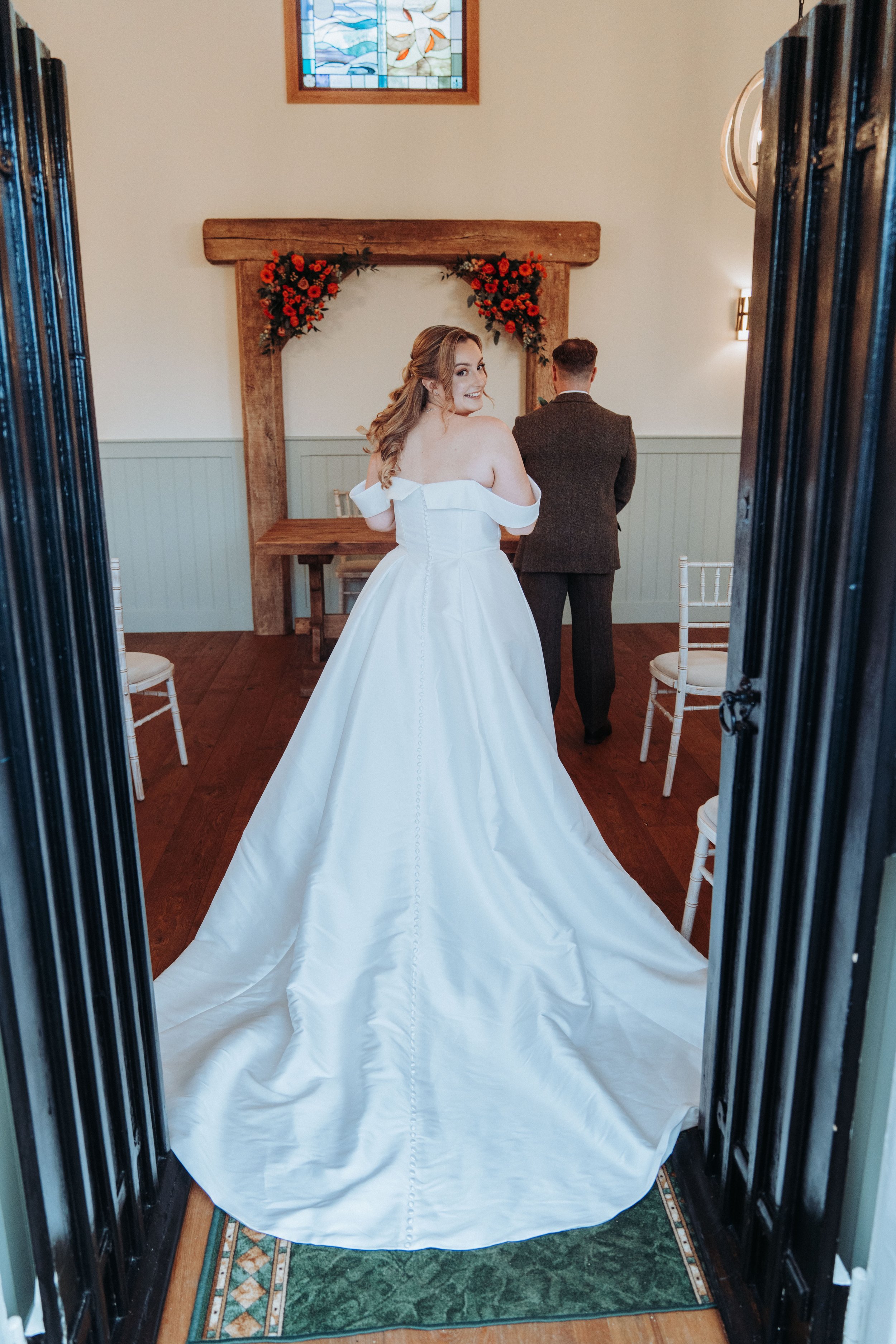 A bride in a white wedding gown standing in a doorway, smiling over her shoulder, with a man in a brown suit in the background, inside a decorated wedding venue.