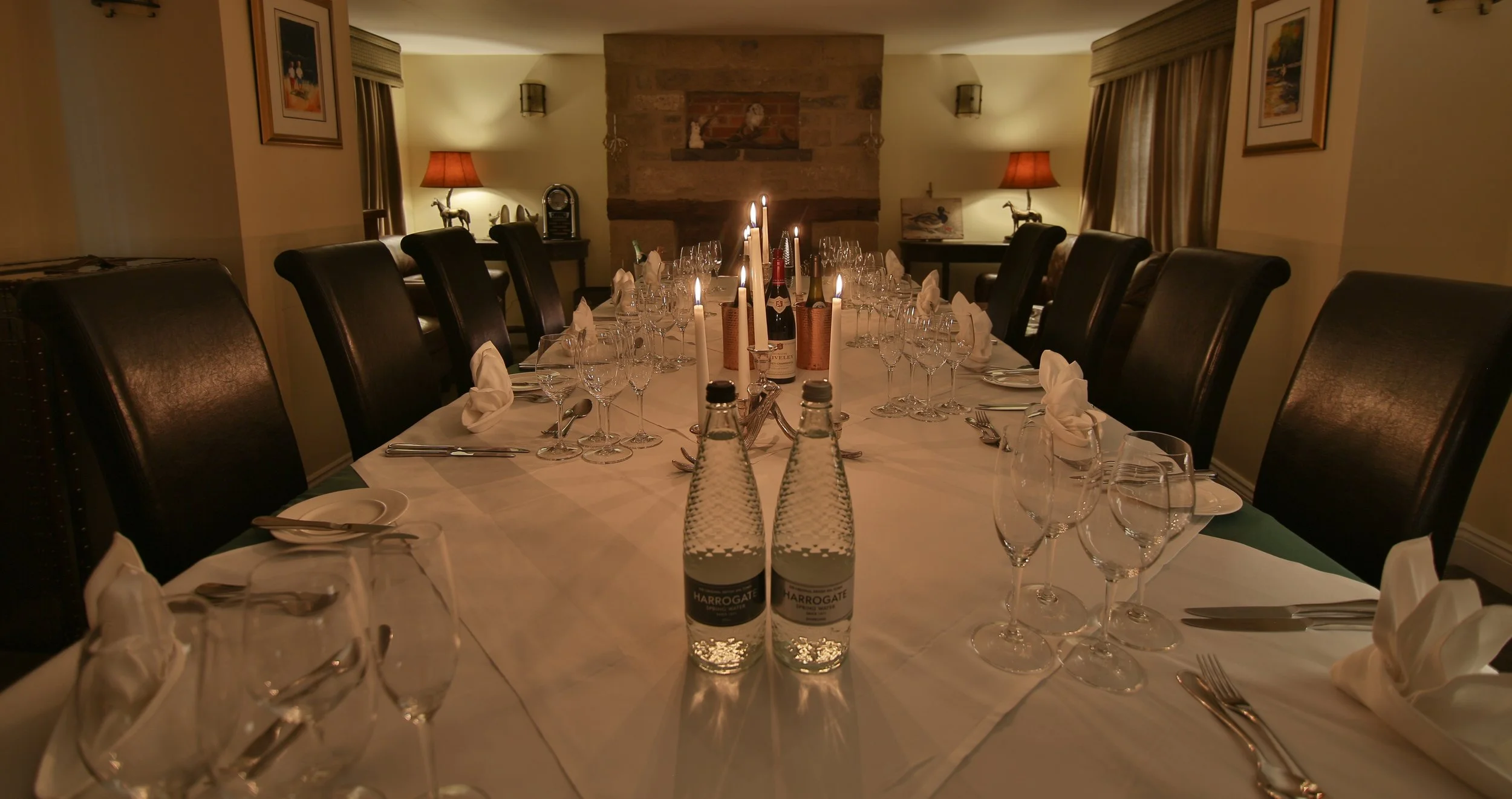 A dining room decorated for a formal dinner with a long table set with glasses, silverware, white napkins, candles, and bottles of water. The room has a brick fireplace, framed art, and curtains, with warm lighting from lamps and candles.