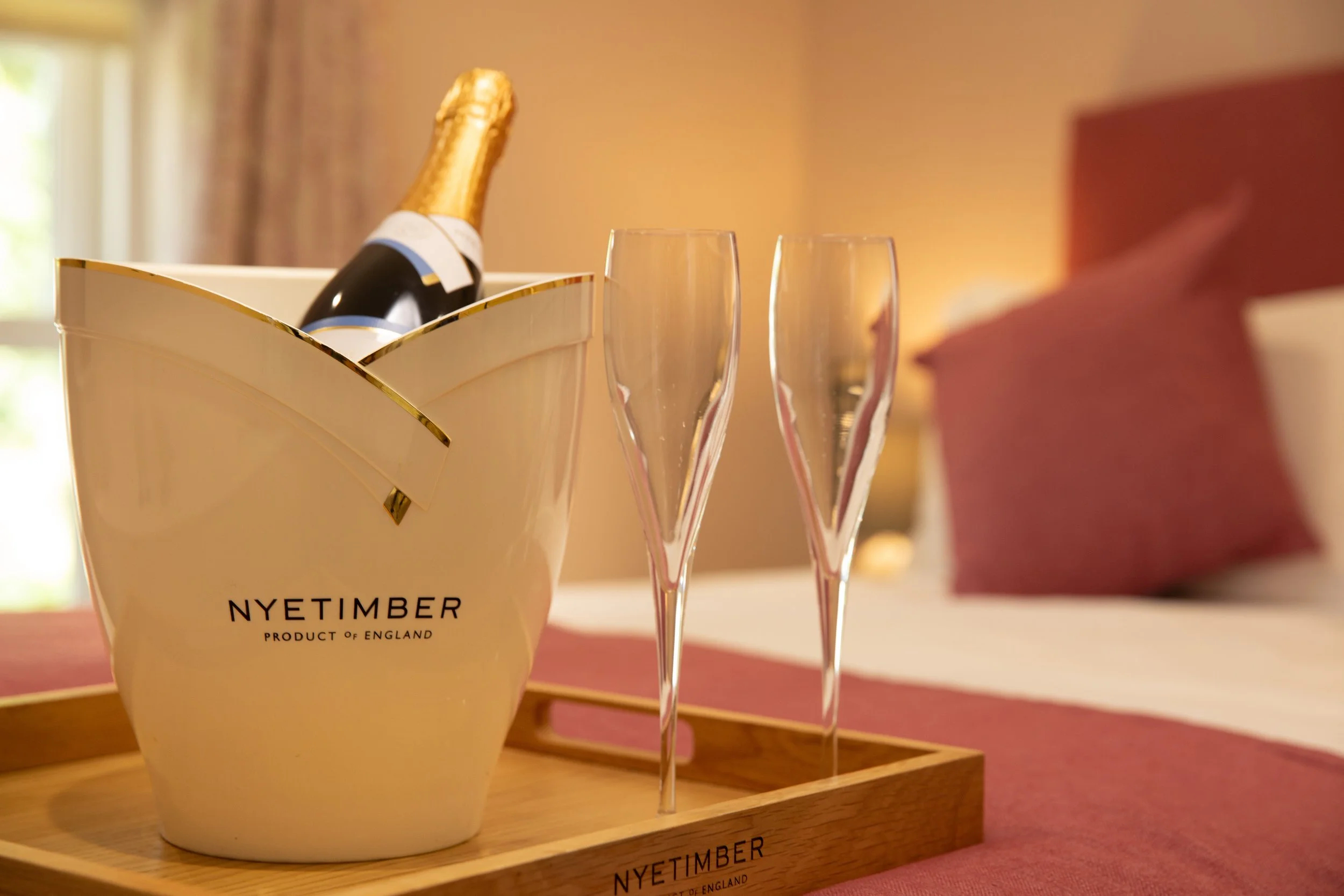 A bottle of champagne in a nyetimber bottle cooler, and two empty champagne flutes on a wooden tray, set on a pink tablecloth in a cozy room with a blurred background.