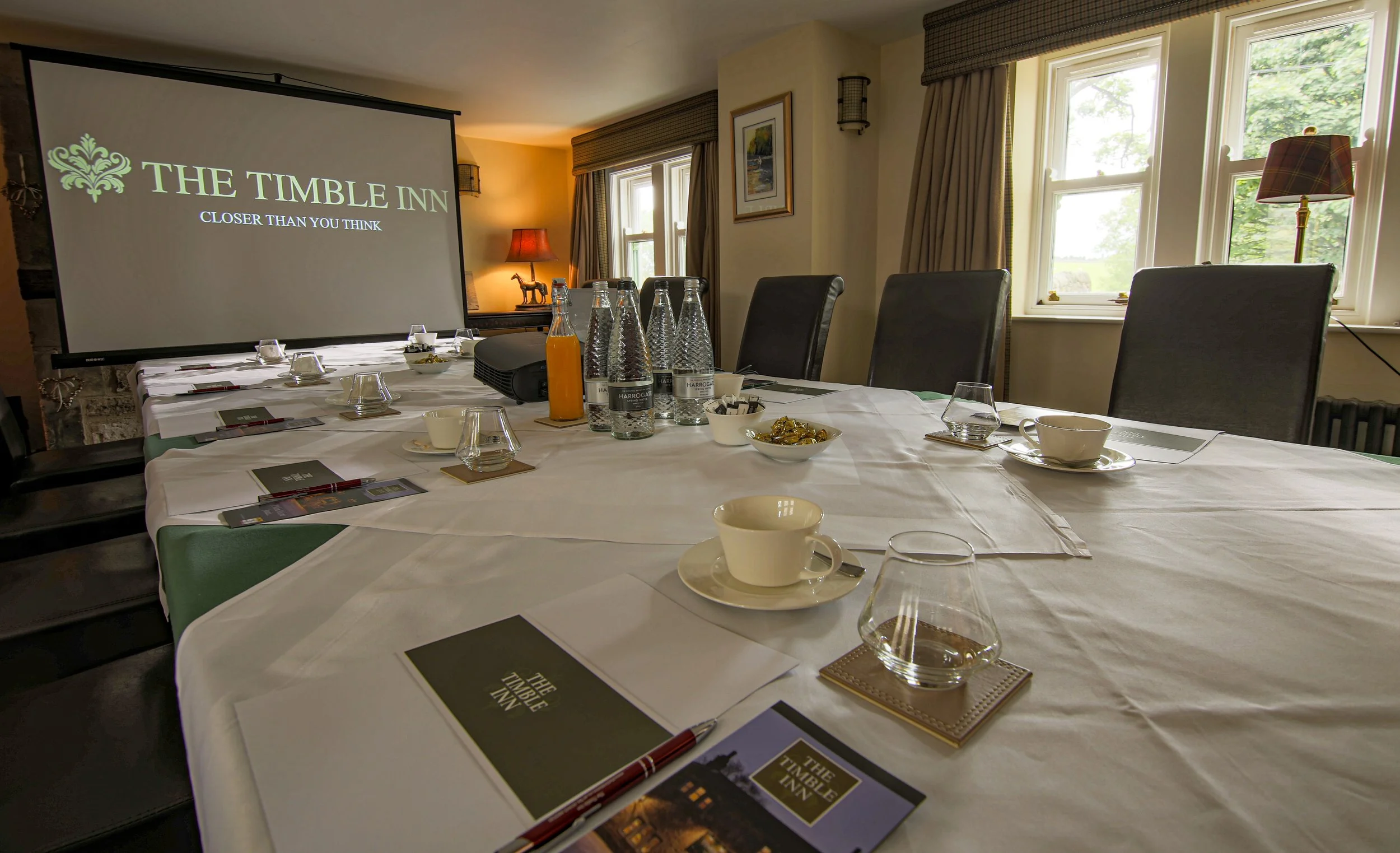 A conference room set up for a meeting with a large table covered in a white tablecloth, water bottles, glasses, cups, and informational pamphlets. There is a large projector screen displaying 'THE TIMBLE INN' along with a slogan. The room has large windows with curtains that let in natural light, and wall-mounted lamps.