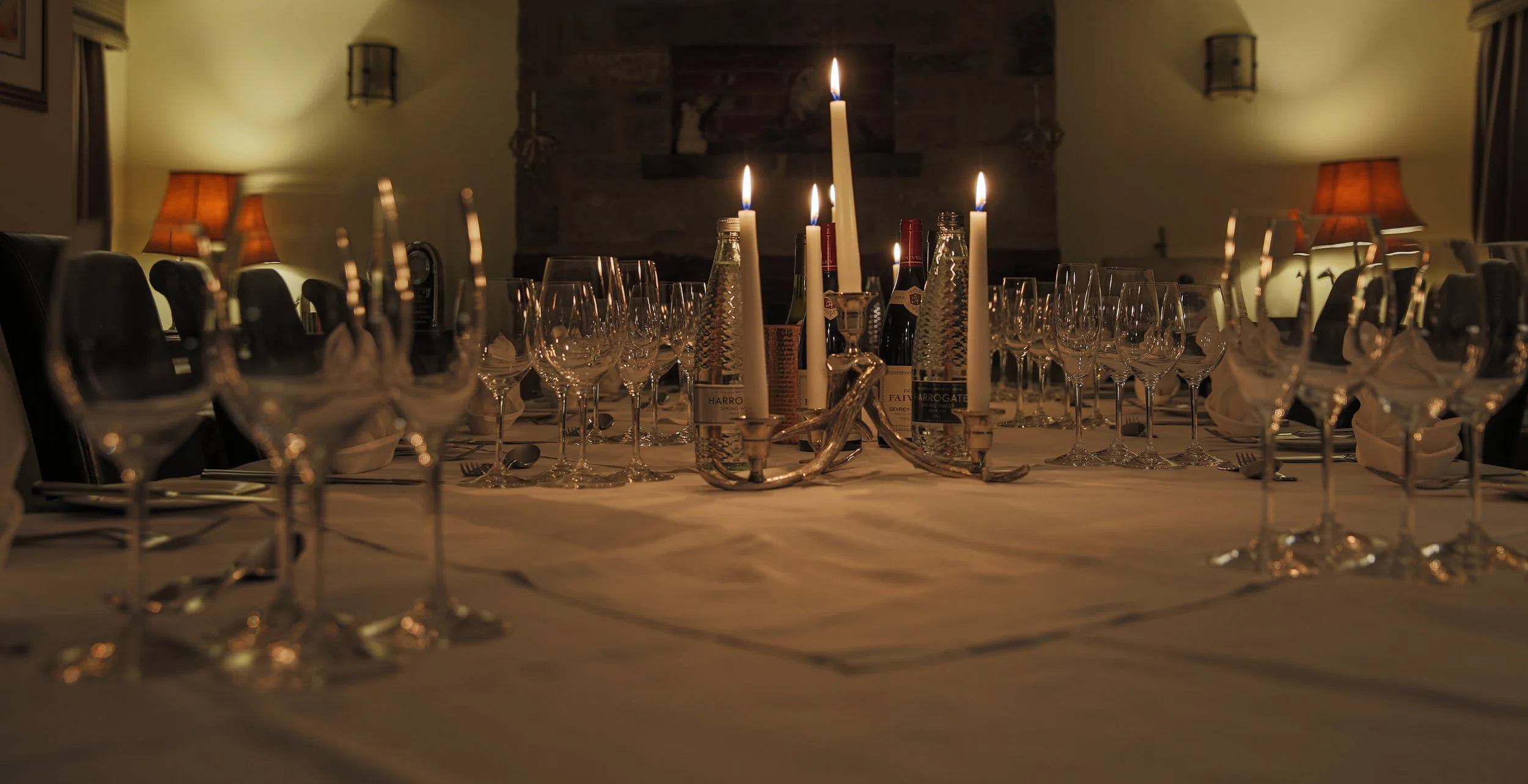 A dimly lit dining table set for a formal meal with multiple empty wine glasses, candles, and bottles of wine, in a cozy room with warm lighting and a brick fireplace.
