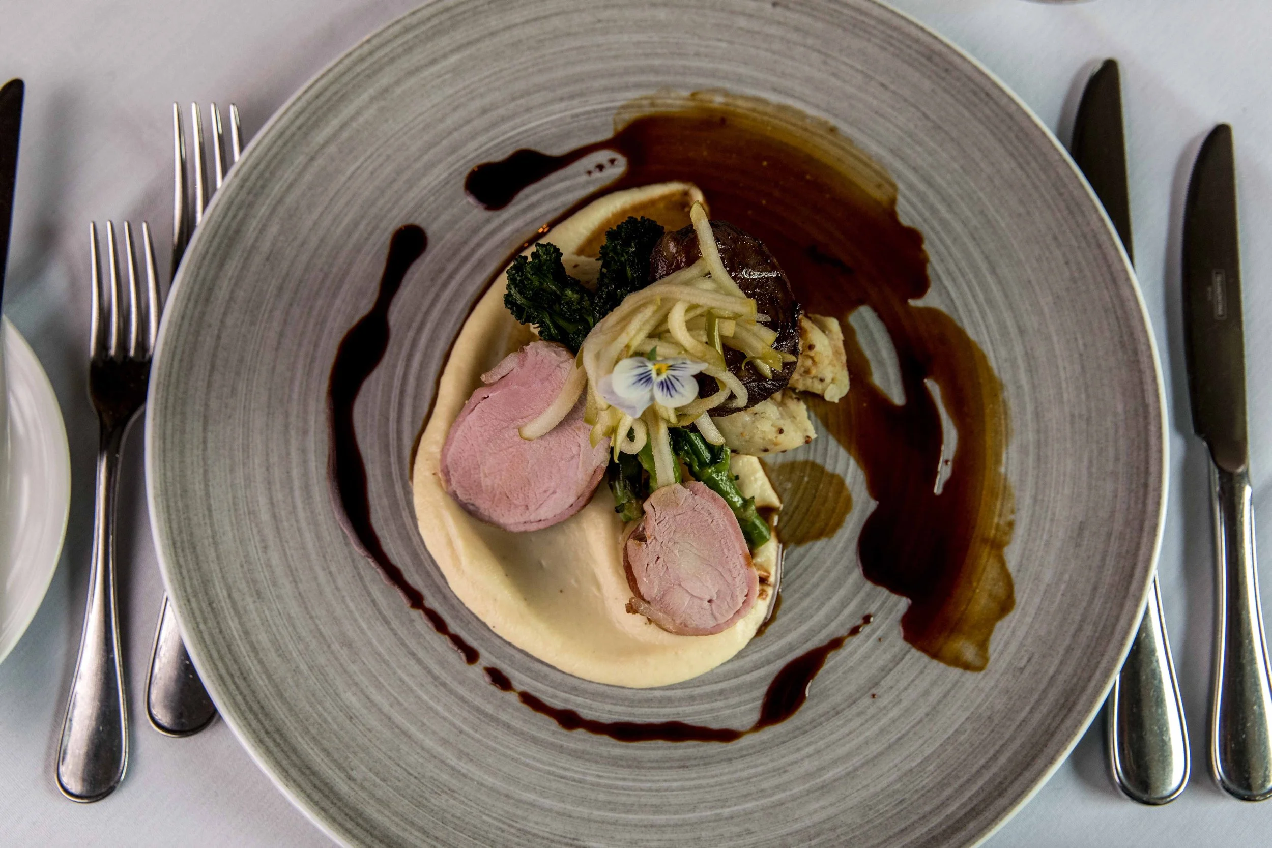 A plated gourmet dish featuring sliced pink meat, mashed potatoes, sautéed greens, a mushroom, and a flower petal garnish, drizzled with dark sauce.