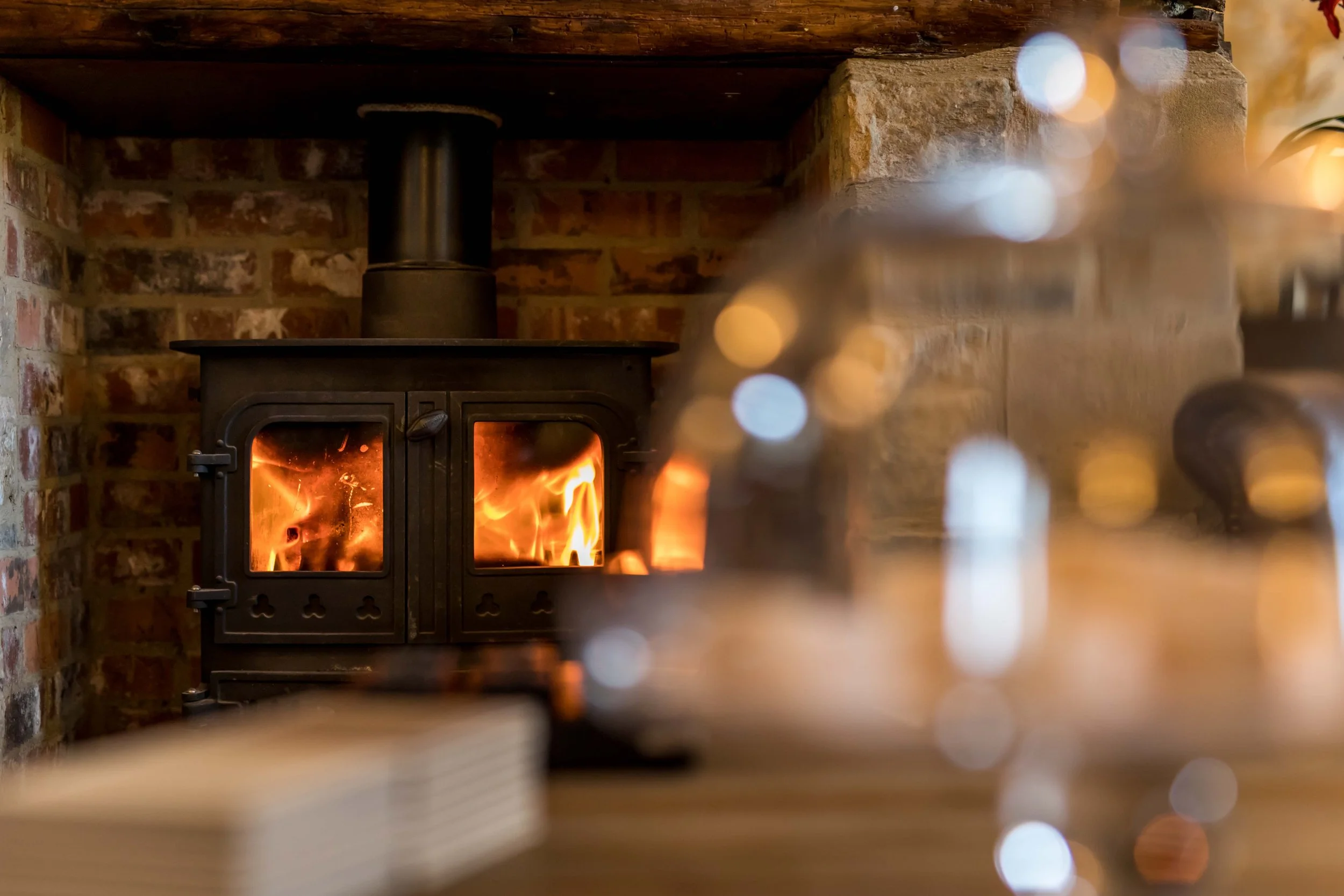 A wood-burning stove with visible flames, set against a brick wall, with a wooden countertop above it.