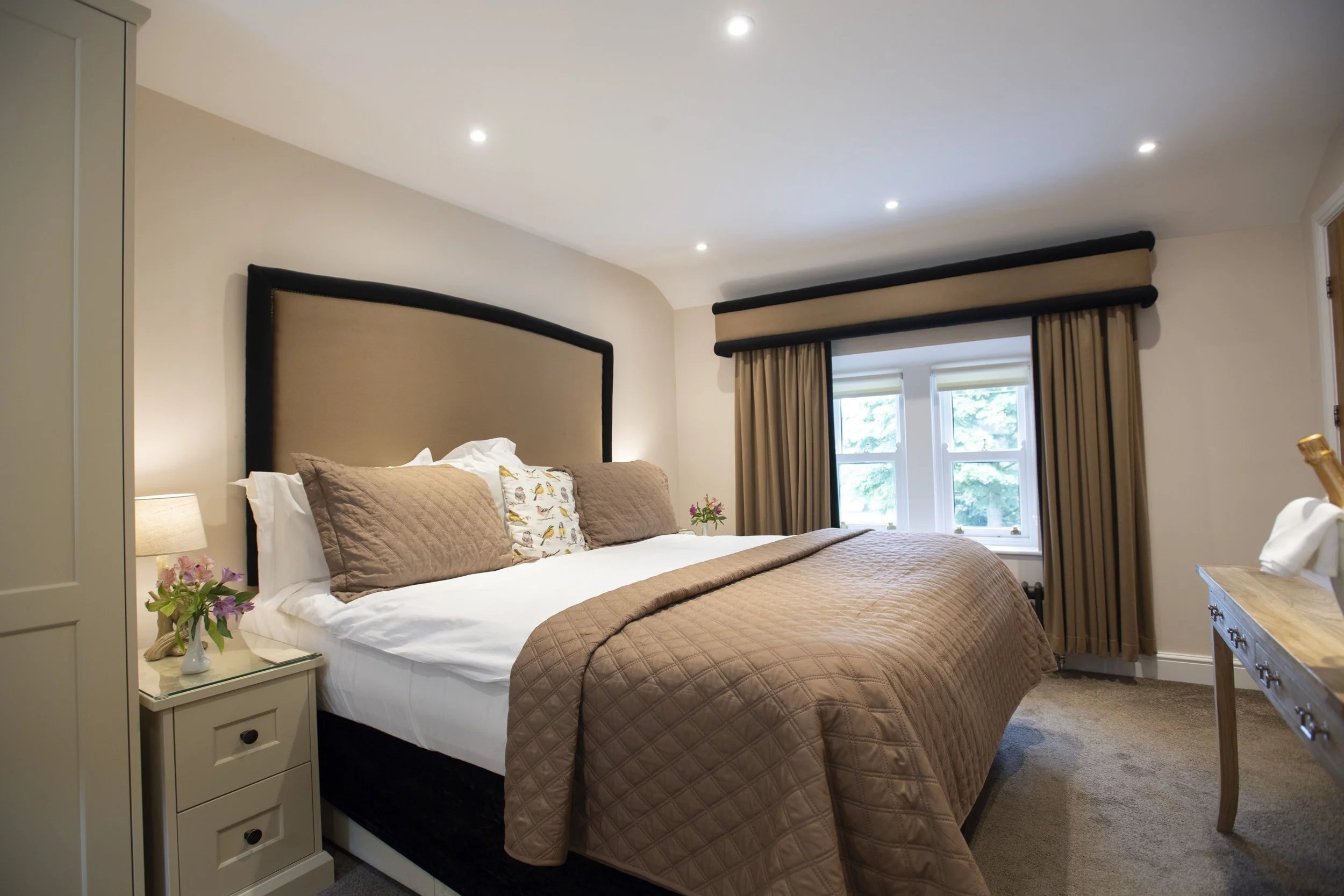 A neatly made bed with brown quilted bedding, multiple pillows, and a decorative pillow with birds, in a cozy bedroom with a window, curtains, and a bedside table with flowers.
