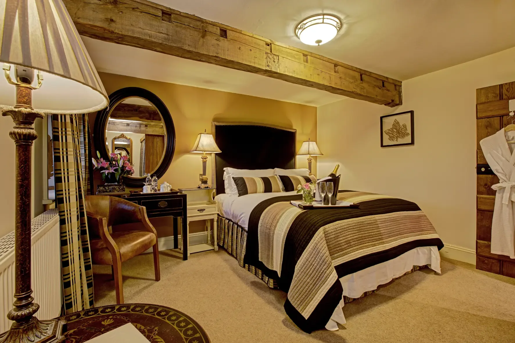 A cozy hotel room with a large bed, side lamps, a mirror, and decorative wall art. The room features warm tones and wooden accents.