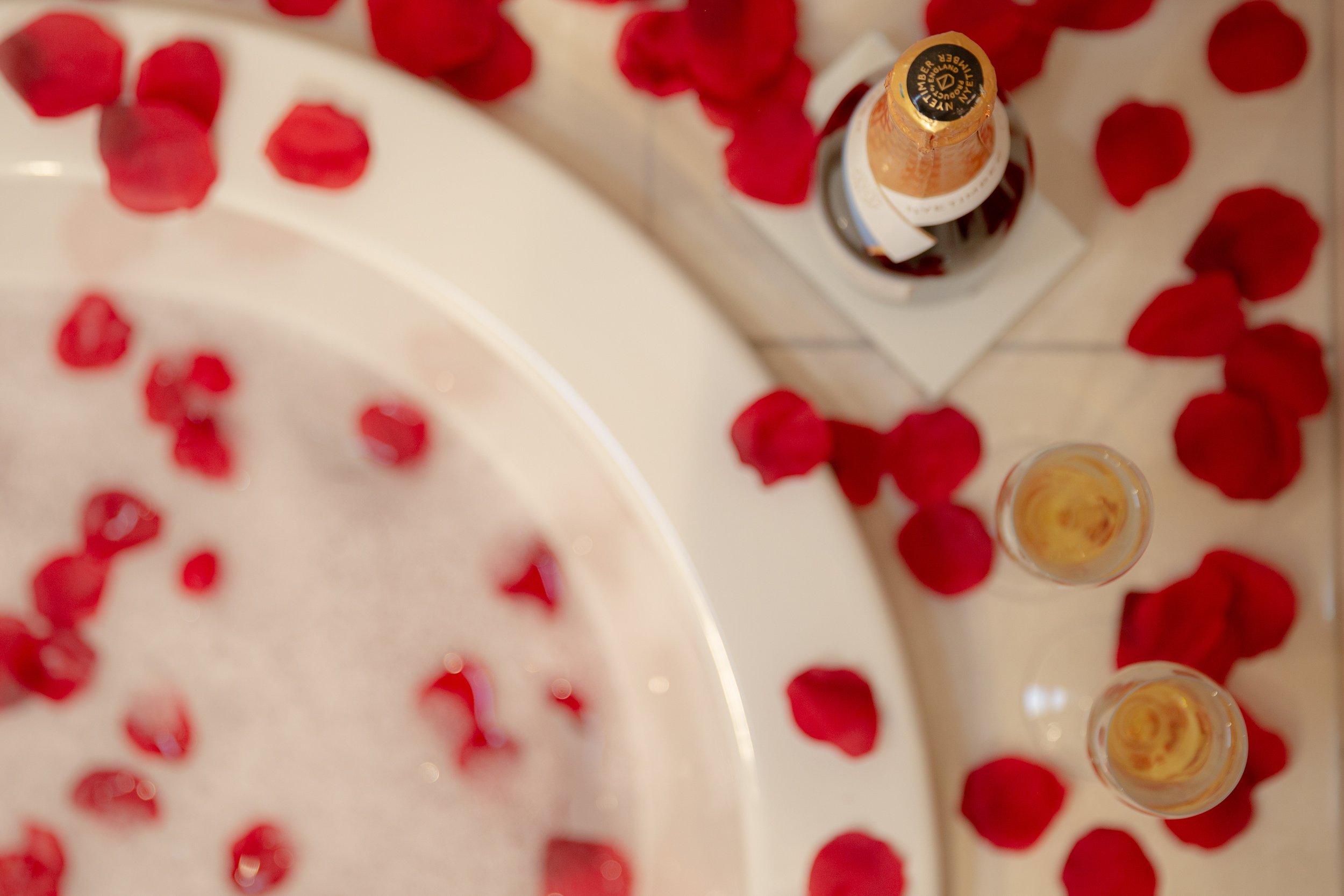 Red rose petals scattered around a bottle of champagne and glasses on a white surface.