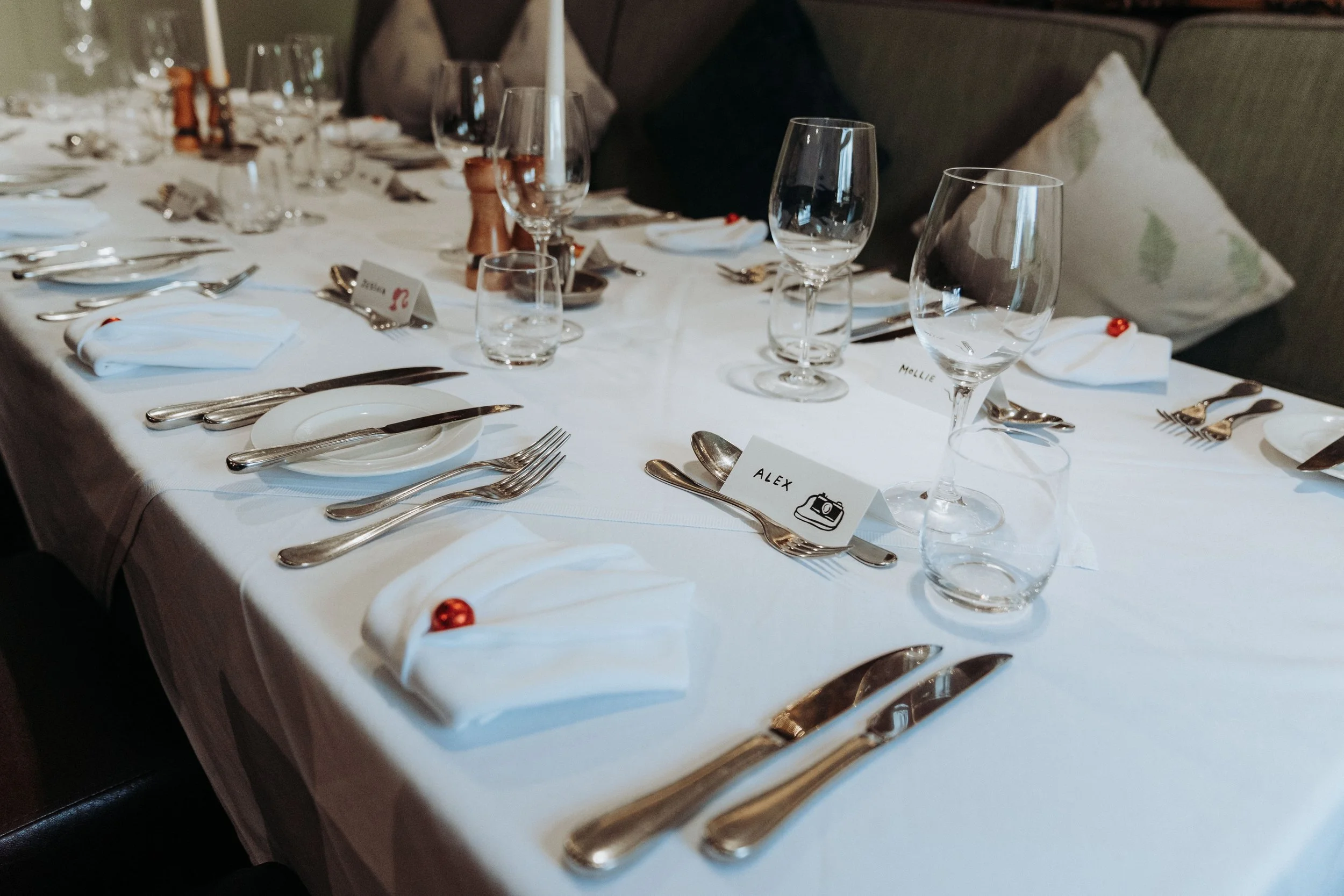 A elegantly set dining table with glassware, silverware, napkins, and place cards for a formal event, featuring white tablecloth and decorative accents.