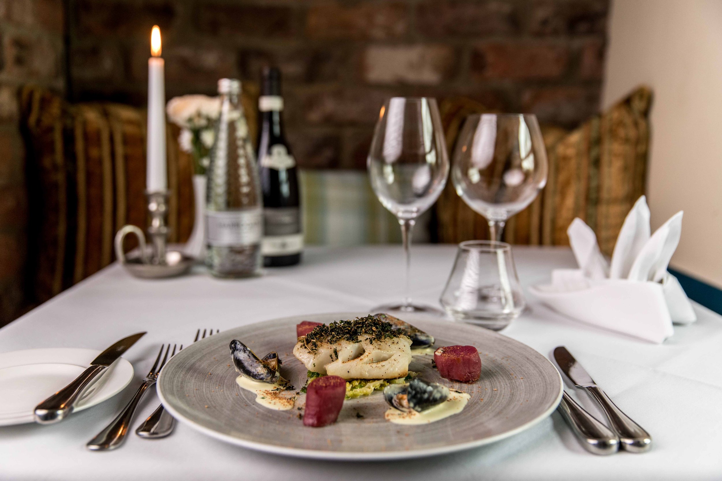 Elegant restaurant table with plated seafood dish, wine glasses, candles, and decorative flowers in the background.