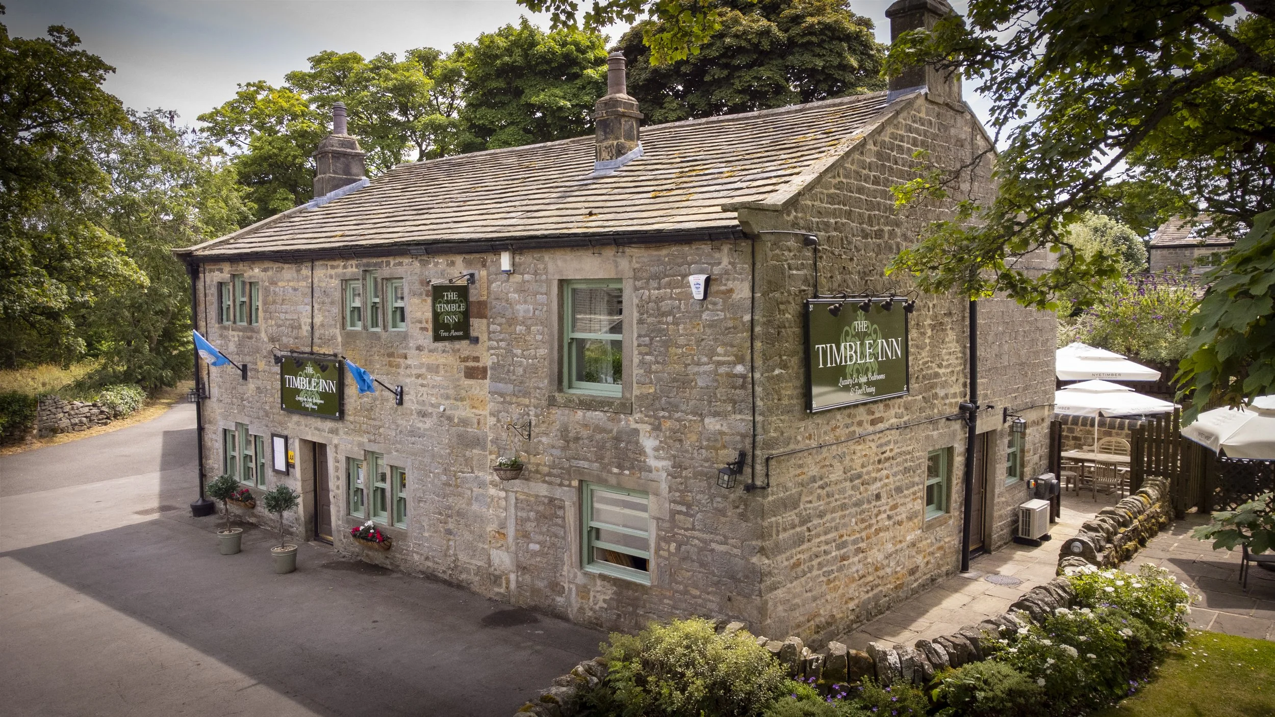 A two-story stone building with signs indicating it is The Timble Inn. The building has multiple green-framed windows, potted plants outside, and outdoor seating with umbrellas. It is surrounded by trees and greenery.