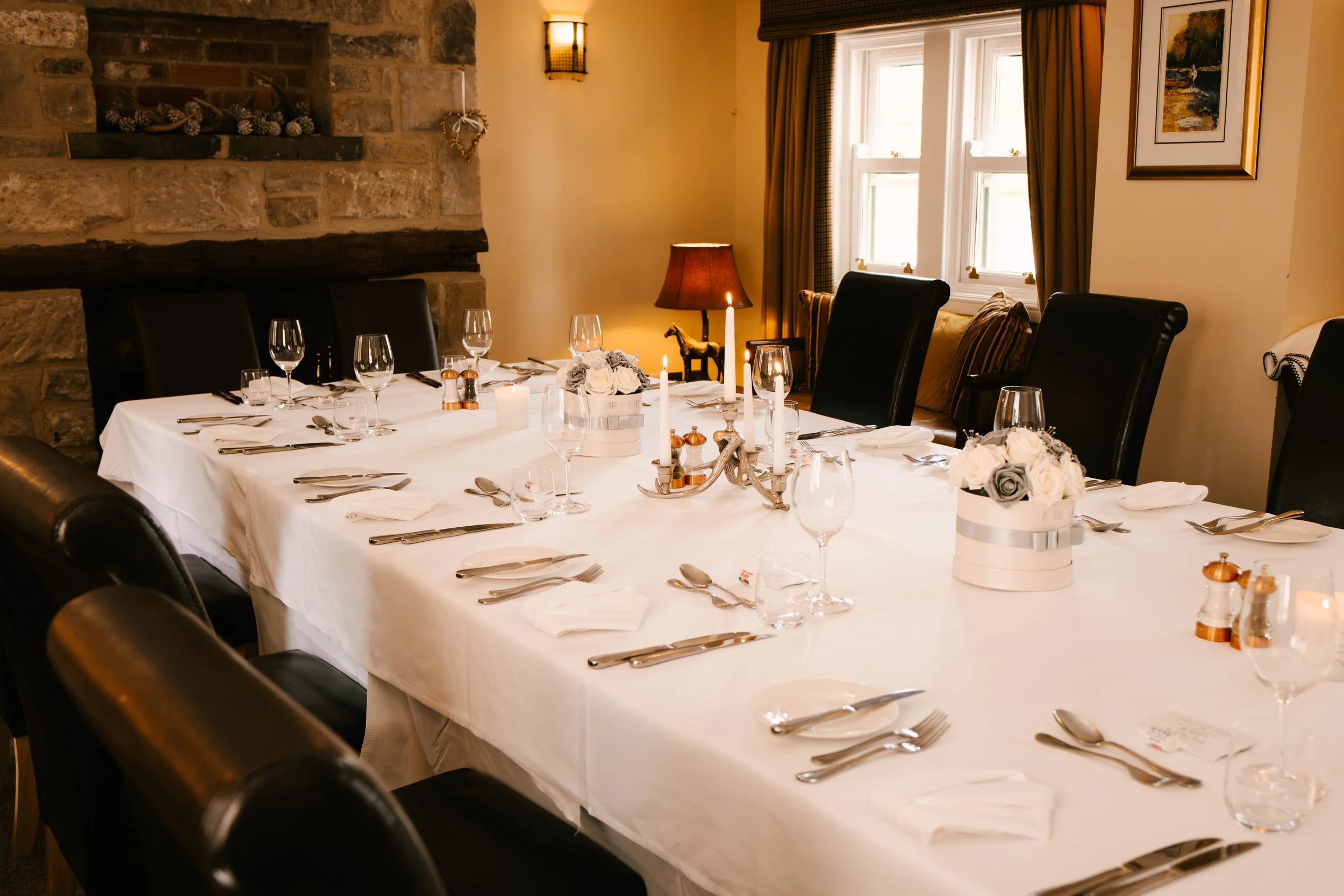 A formal dining room with a long table covered with a white tablecloth, set for a meal with plates, silverware, and glasses. There are candles and flower centerpieces on the table. The room has a stone fireplace on one wall, framed artwork, and windows with yellow curtains. A side table has a lamp and decorative items.