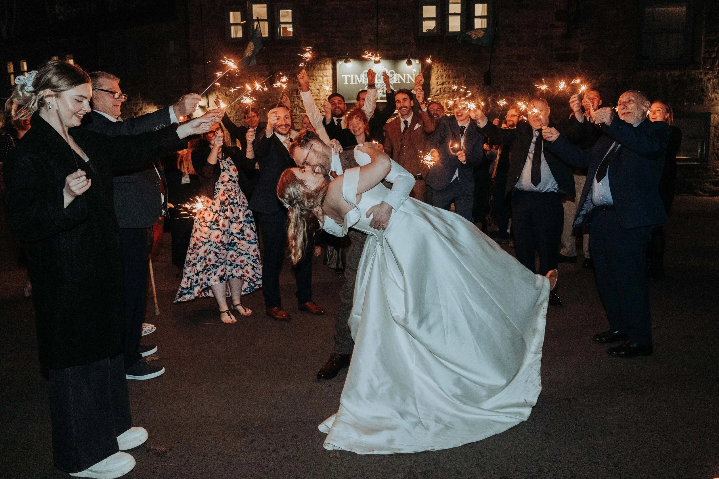 A group of people celebrating at night, holding sparklers. A newly married couple is in the center, with the bride kissing the groom. The setting appears to be a festive event, possibly a wedding reception, outside with a sign in the background that reads 'TIME INN'.