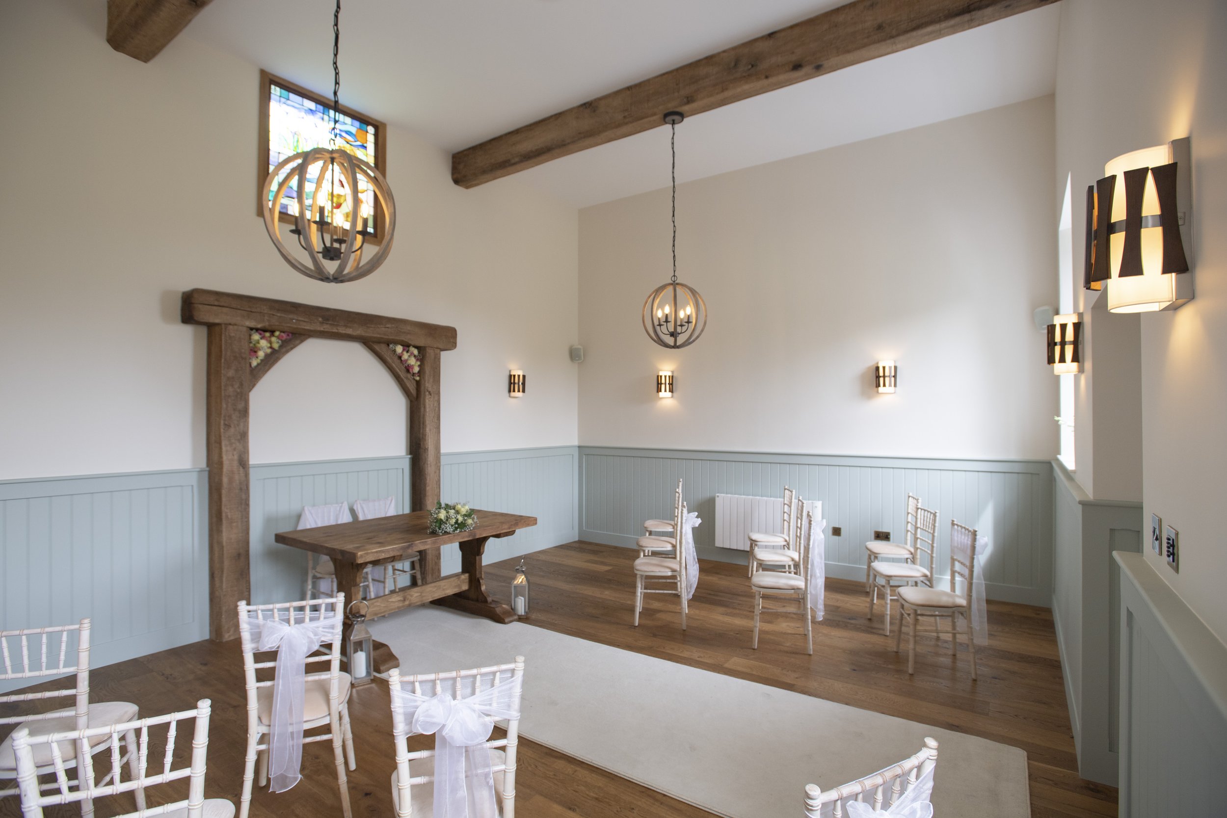 A decorated wedding ceremony room with white and light blue walls, wooden beams, a stained glass window, chandeliers, wall sconces, and chairs with white covers and bows.