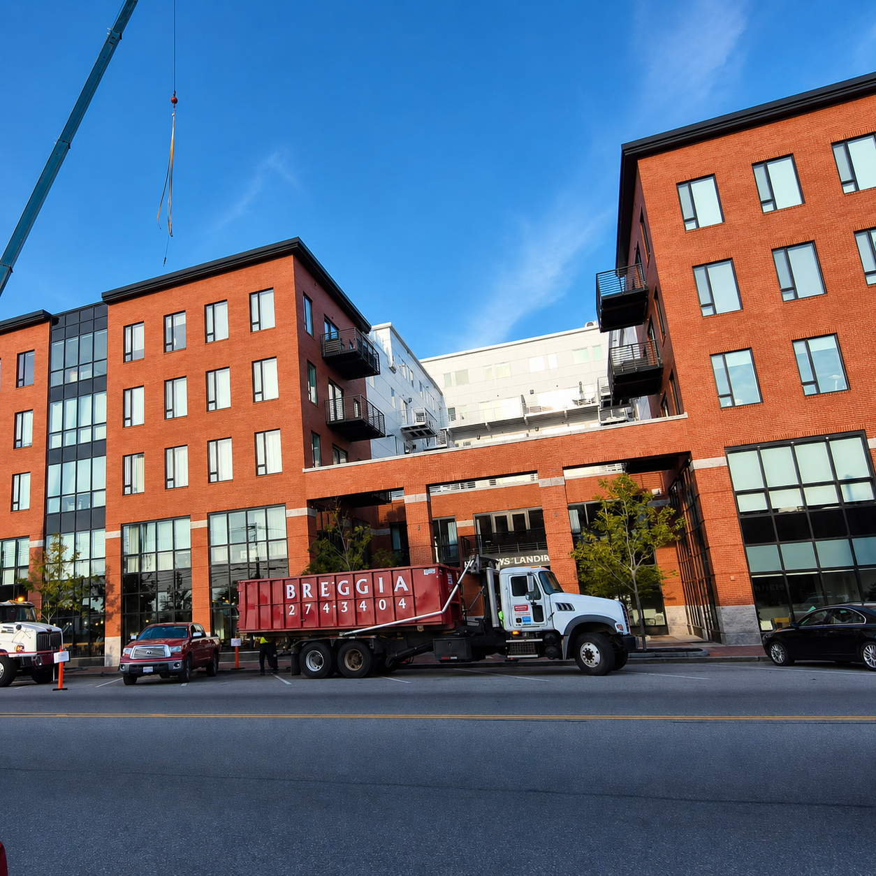 City street with a modern red brick apartment building, cars parked, and a construction truck on the road.