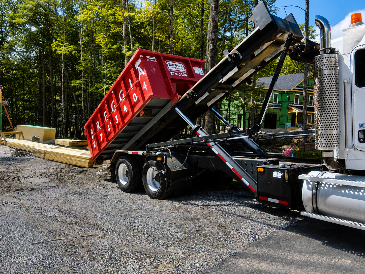 A construction truck with a tilted red container lifting wood planks at a building site surrounded by trees and under construction houses.