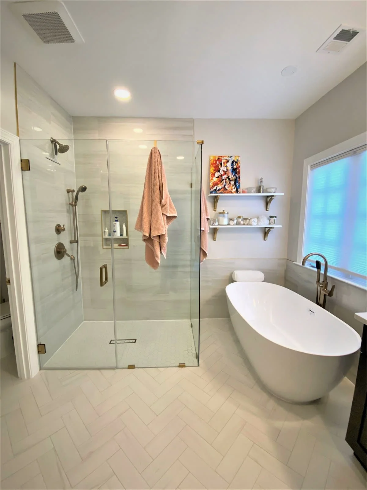 Modern bathroom with a glass-enclosed shower area, a white freestanding bathtub, two pink towels, wall-mounted shelves with decor, a window with blinds, and a painting on the wall.