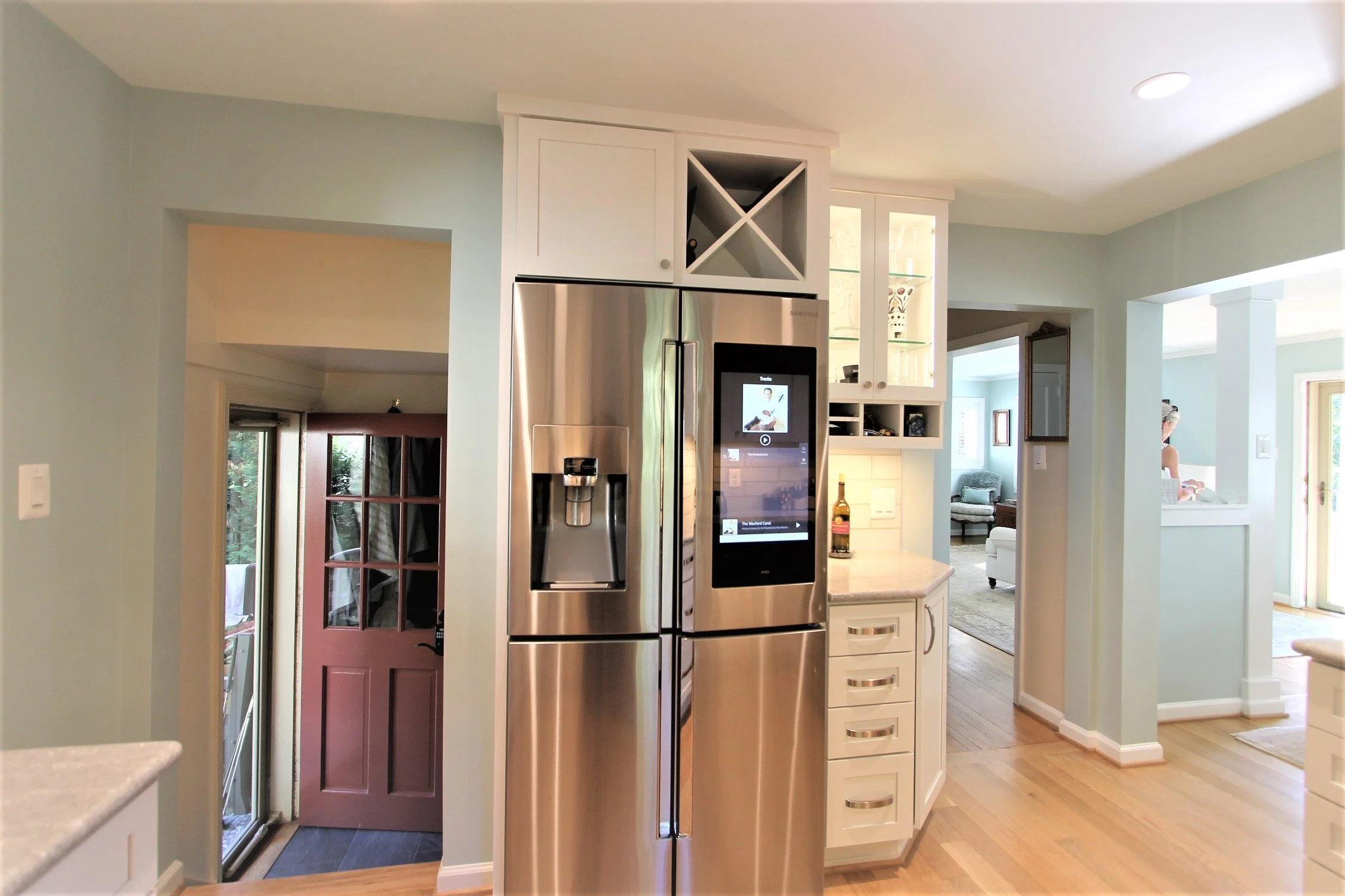 The interior of a house showing a stainless steel refrigerator next to a white kitchen cabinet with an electronic display panel. There is a purple door leading outside and a living room visible in the background.