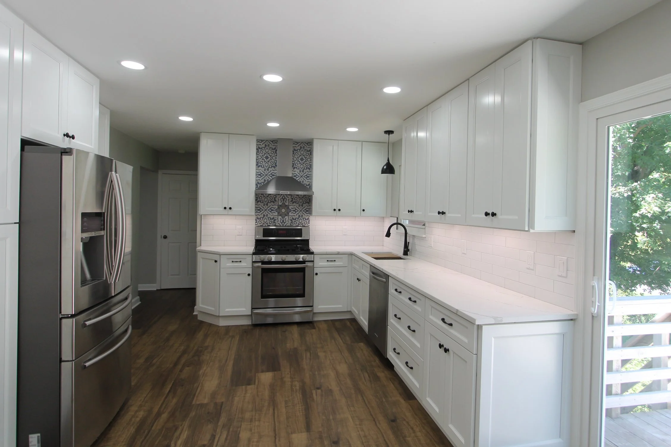 Modern white kitchen with stainless steel appliances, dark wood flooring, and large window showing green trees outside.