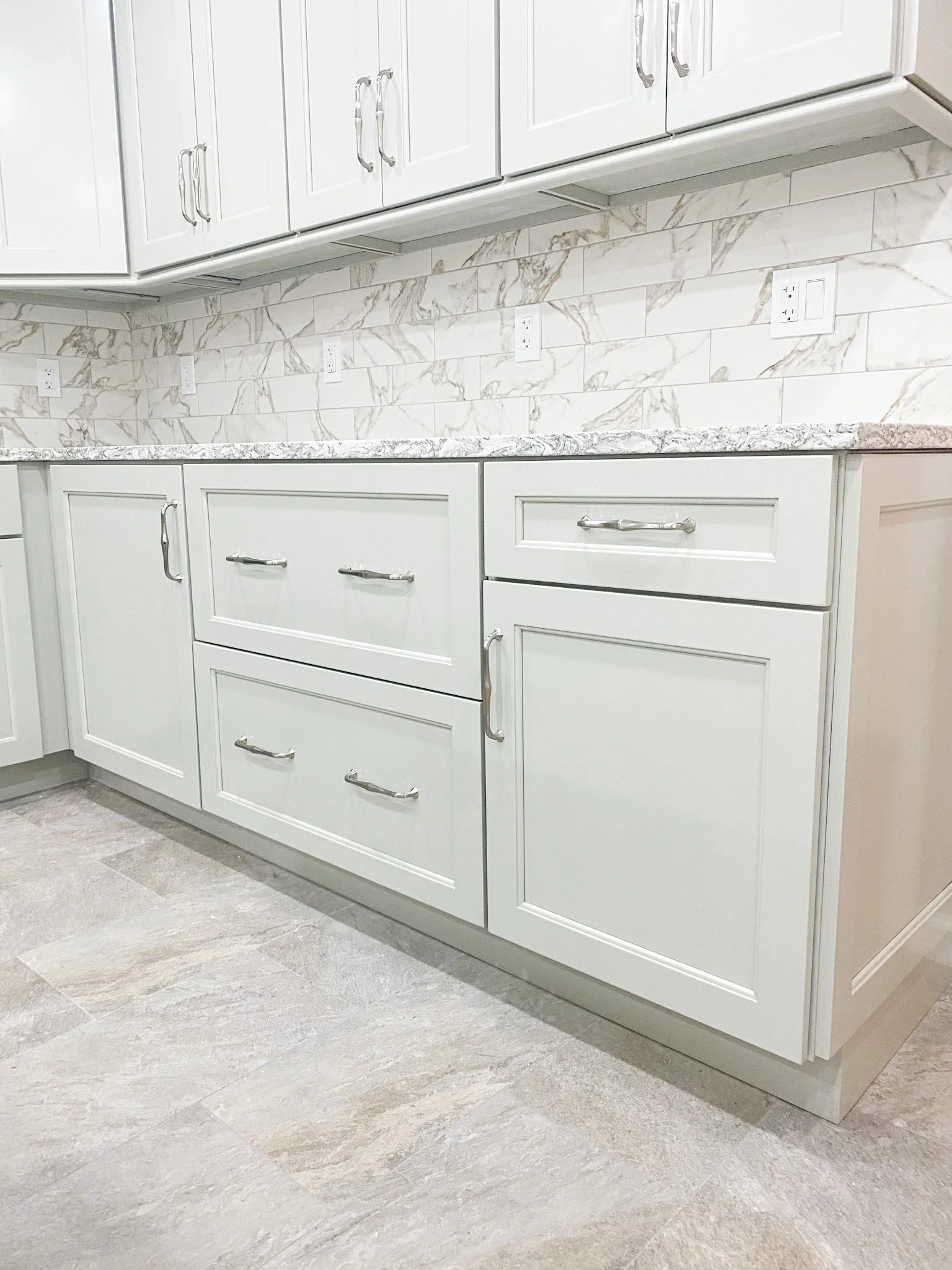 Kitchen with white cabinetry, marble-like countertops, marble backsplash, electrical outlets, and beige tiled floor.