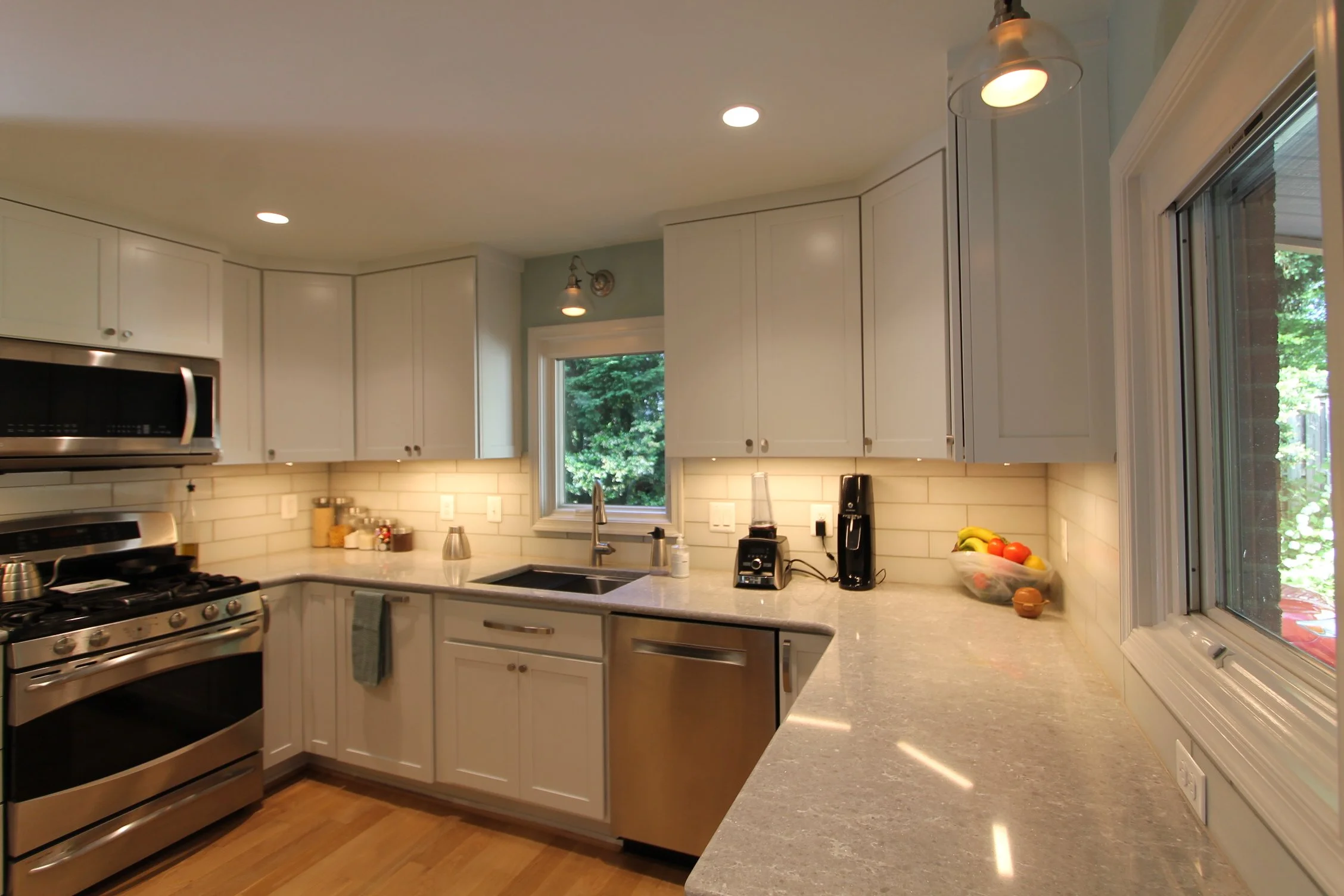 Kitchen with white cabinets, stainless steel oven and microwave, beige countertops, window above sink, and various small appliances on the counter.