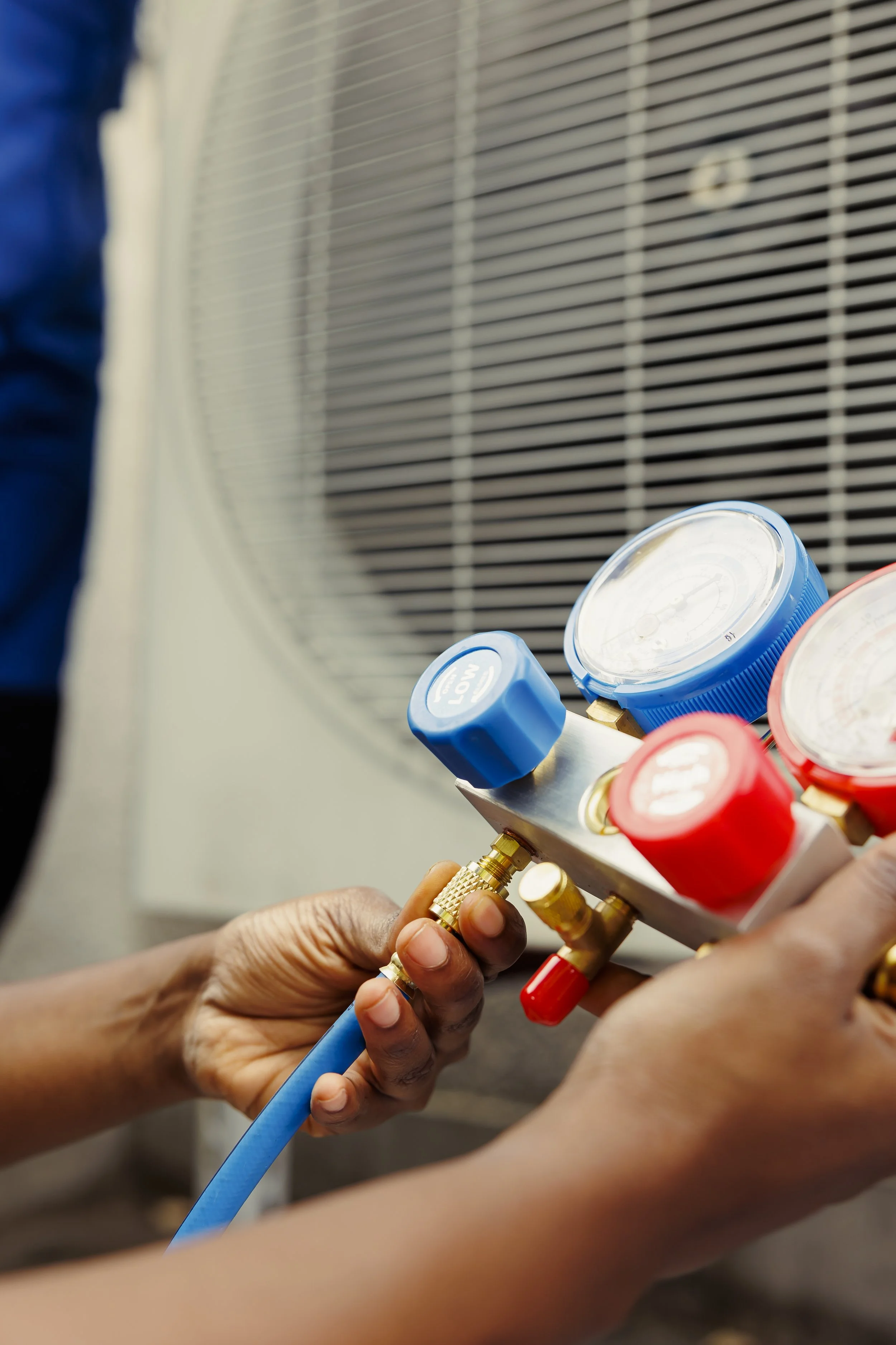 Two people are connecting hoses to a manifold, likely for refrigerant or air conditioning system servicing. One person's hand is holding a blue hose, while a second hand is adjusting the connection to the manifold.
