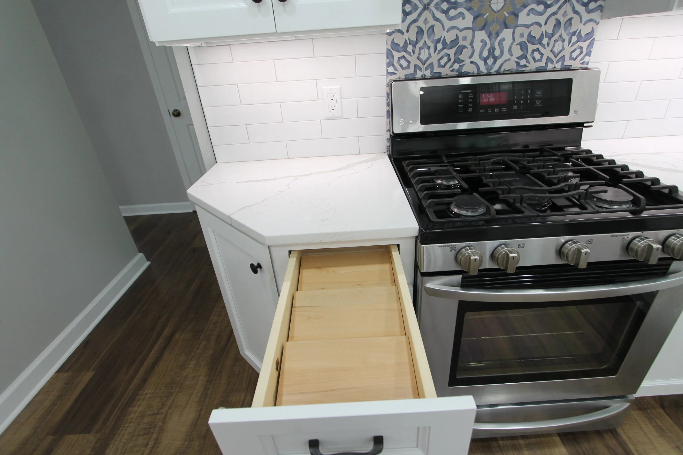 A kitchen with a white countertop, white cabinets, and a gas stove. There is a decorative tiled backsplash behind the stove with blue and white patterns. An electrical outlet is visible on the backsplash, and a small drawer in a white cabinet is pull