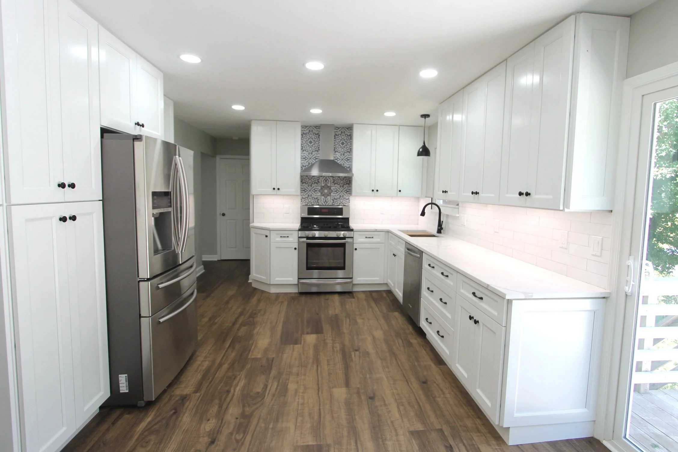 A modern kitchen with white cabinets, a stainless steel refrigerator, a stove, black faucet, and a window with a view of trees outside.
