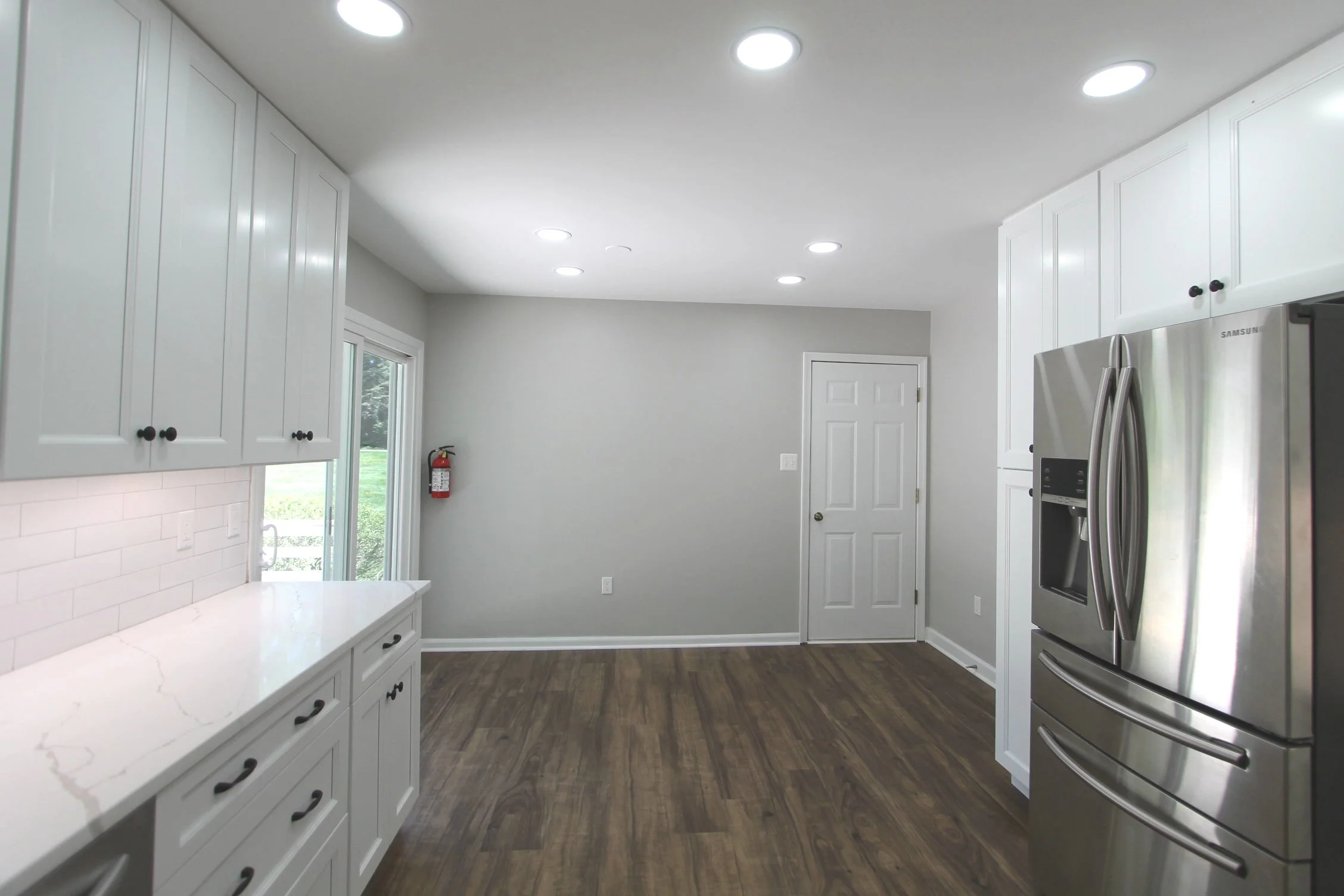 Modern kitchen with white cabinets, a white marble countertop, a stainless steel side-by-side refrigerator with water dispenser, hardwood flooring, a window with outdoor view, and a door leading to another room.