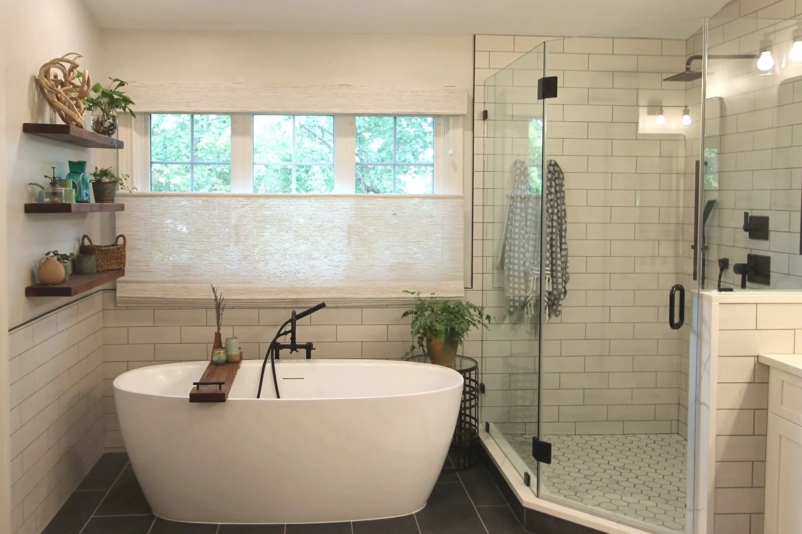 Modern bathroom with a white freestanding bathtub, glass shower enclosure with black fixtures, a window with a textured shade, and decorative shelves with plants and ceramics.