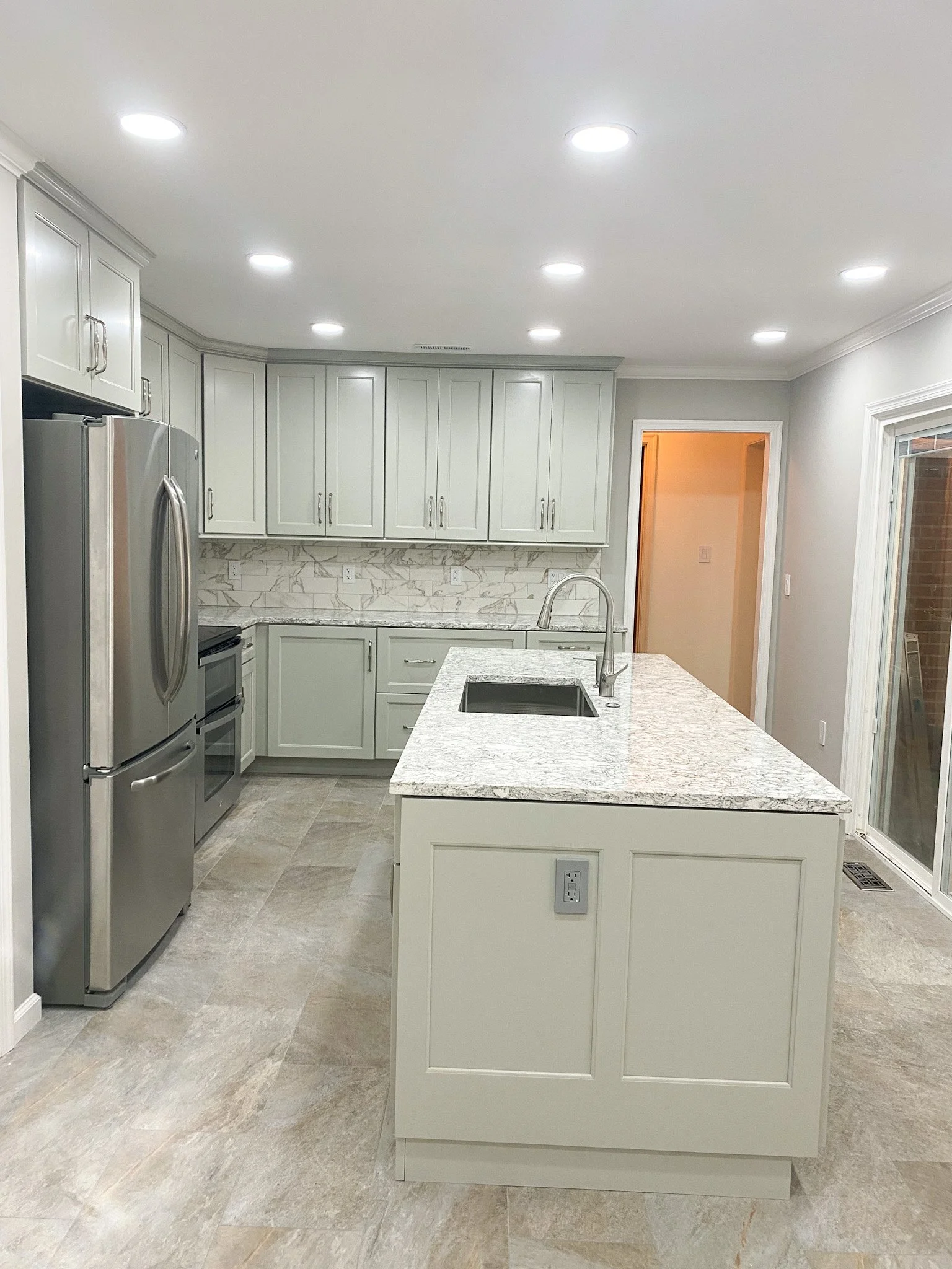 Modern kitchen with white cabinets, marble backsplash, granite countertop island, stainless steel refrigerator, and tile flooring.