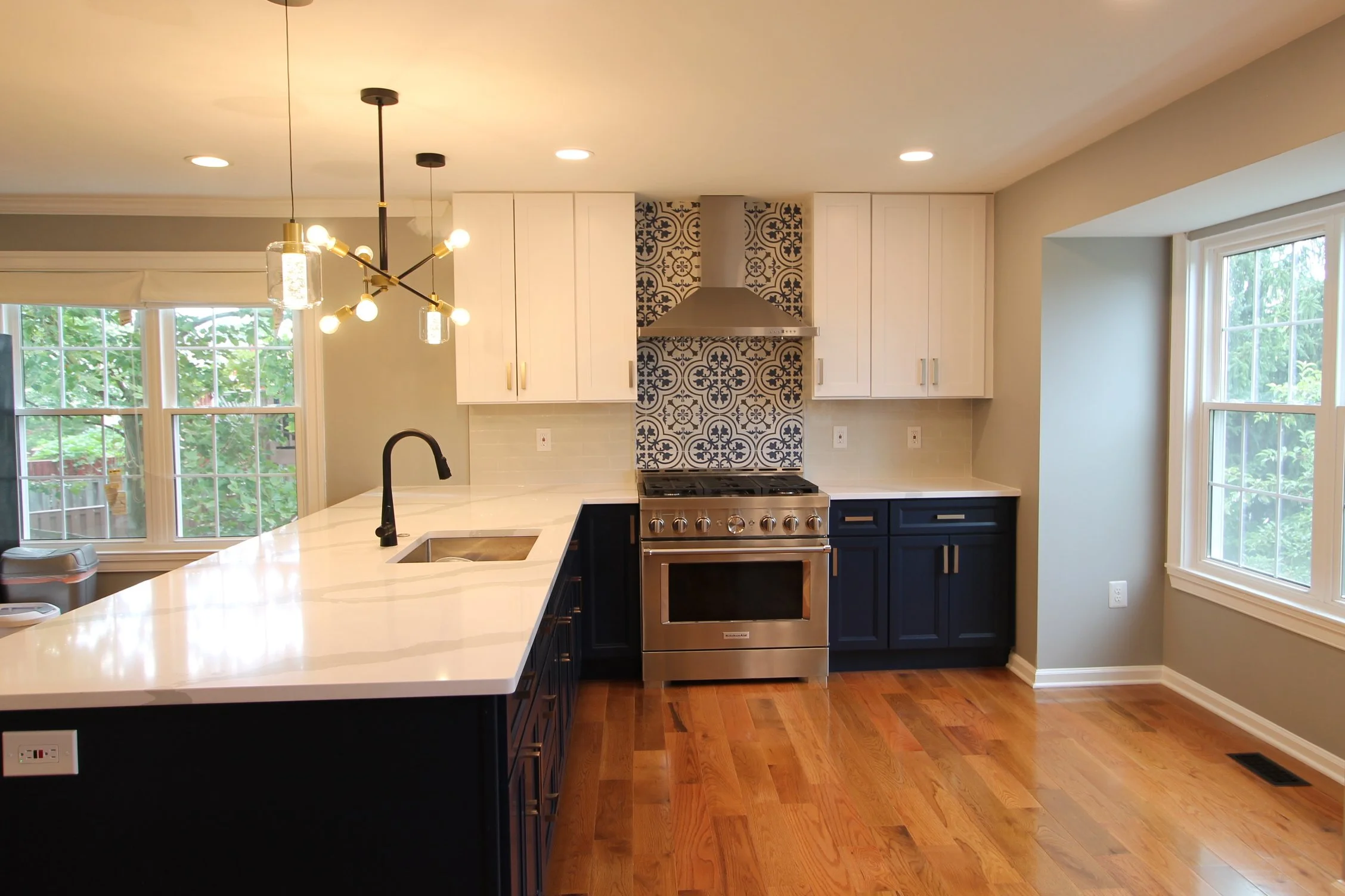 A modern kitchen with white upper cabinets, dark lower cabinets, a stainless steel stove, a black kitchen faucet, and a white countertop island. There are large windows with views of greenery outside, and a decorative tile backsplash behind the stove