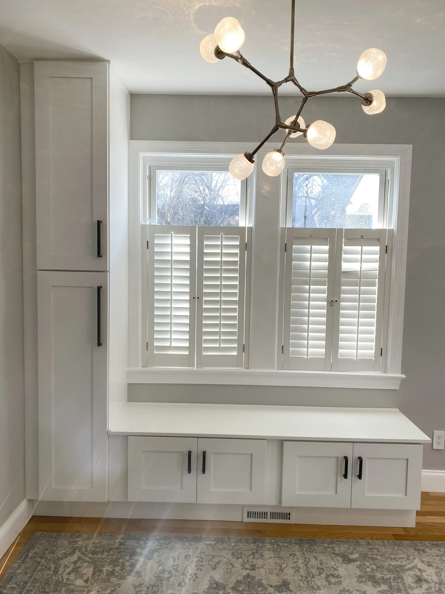 White built-in storage cabinet with cabinets, drawers, and a countertop, positioned in front of windows with white plantation shutters, under a modern chandelier with multiple globe lights, with a patterned rug and hardwood floor beneath.