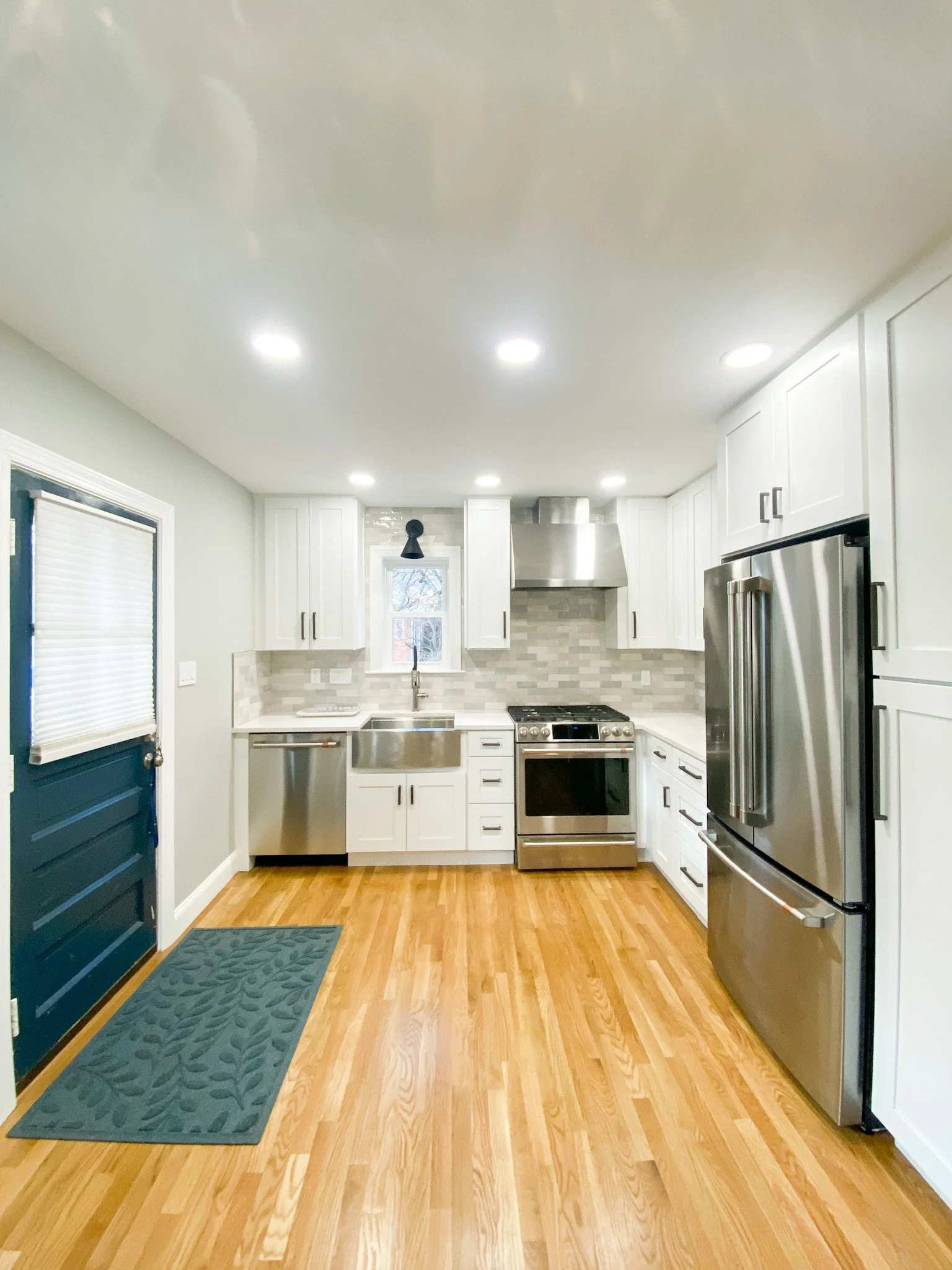 A modern kitchen with white cabinets, stainless steel appliances including a refrigerator and stove, and a brick-style backsplash. There is wood flooring and a small window above the sink. The ceiling has multiple recessed lights.