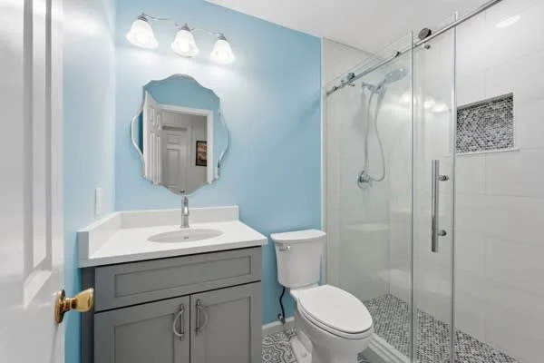 Bathroom with light blue walls, a white vanity with a grey cabinet, a round mirror, a toilet, and a glass shower enclosure with a mosaic tile on the shower floor and a small recessed niche.