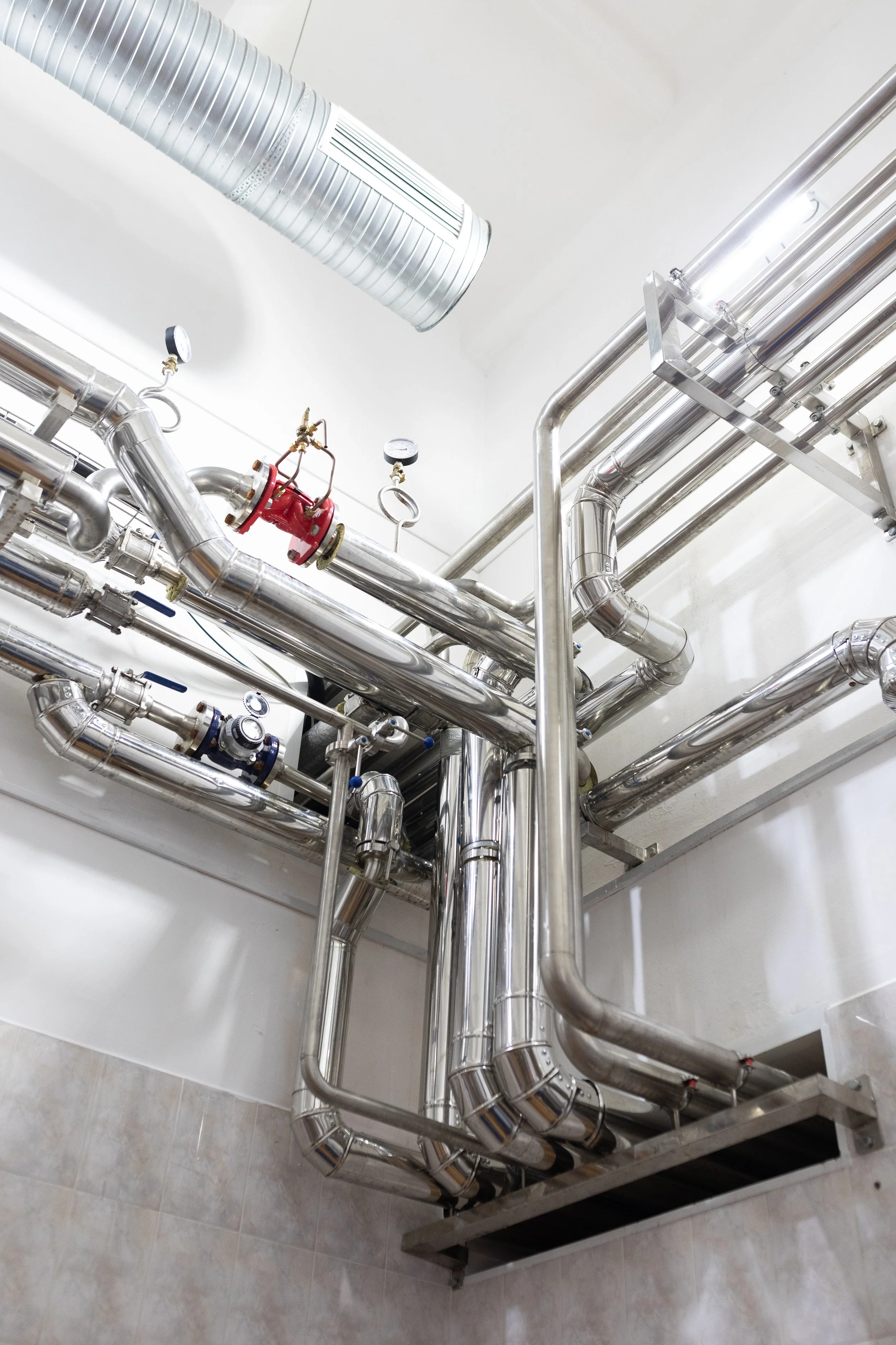 A network of stainless steel pipes with gauges, valves, and insulated sections, mounted on a wall and supported by a frame, in an industrial or mechanical setting.