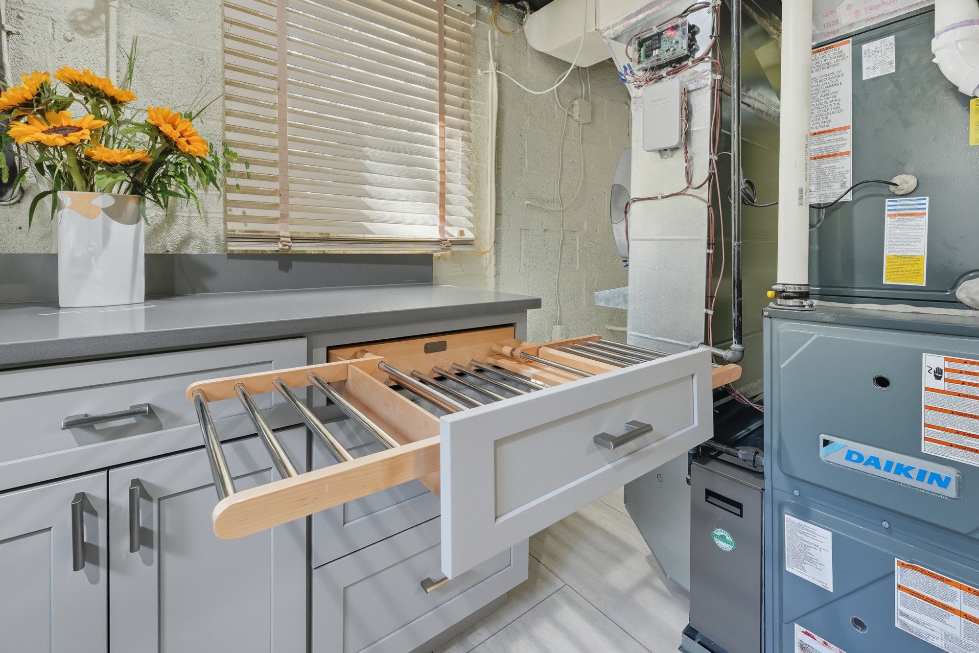 An empty kitchen drawer partially pulled out from white cabinets, with a vase of orange sunflowers on a gray countertop. A window with beige blinds and some exposed pipes and a furnace are visible in the background.