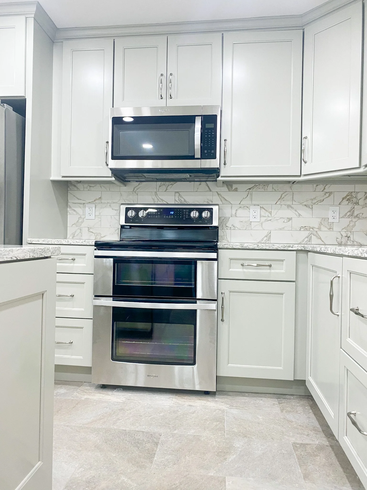 Kitchen with white cabinets, marble backsplash, stainless steel microwave and oven, and light-colored tile floor.