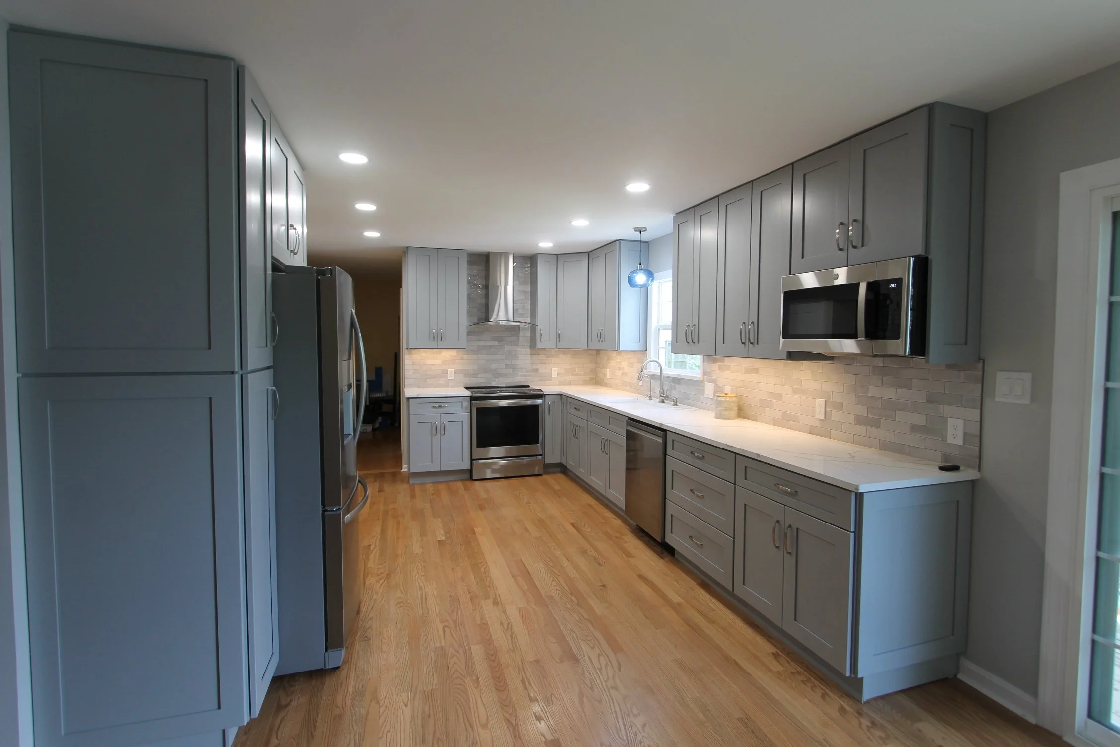 Modern kitchen with gray cabinets, stainless steel appliances, hardwood floors, and a light-colored tile backsplash. Recessed ceiling lights and a small blue pendant light near a window.