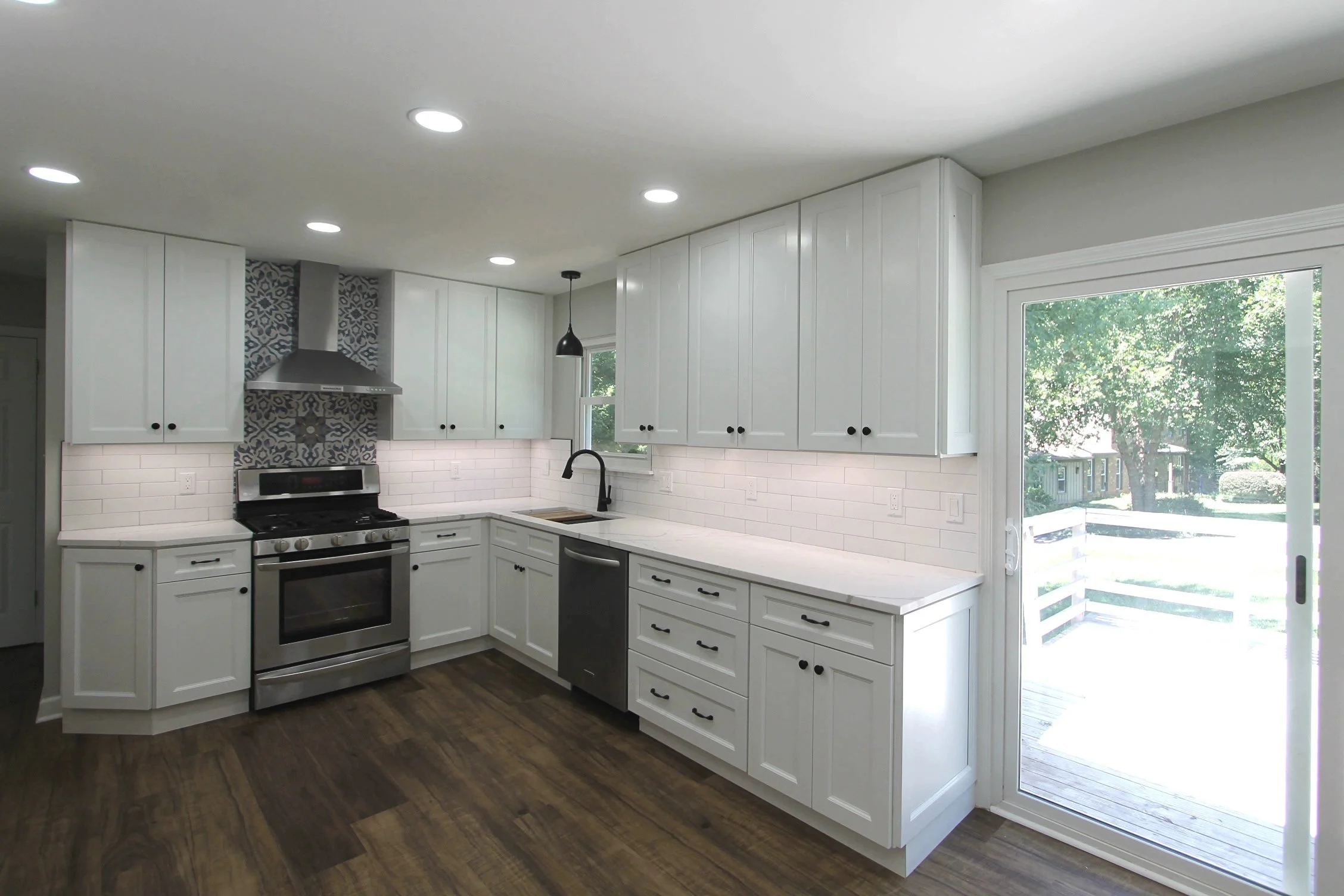 Modern kitchen with white cabinets, black hardware, stainless steel appliances including an oven, vent hood, and dishwasher, wooden flooring, a black pendant light, white subway tile backsplash, and a sliding glass door leading outside.