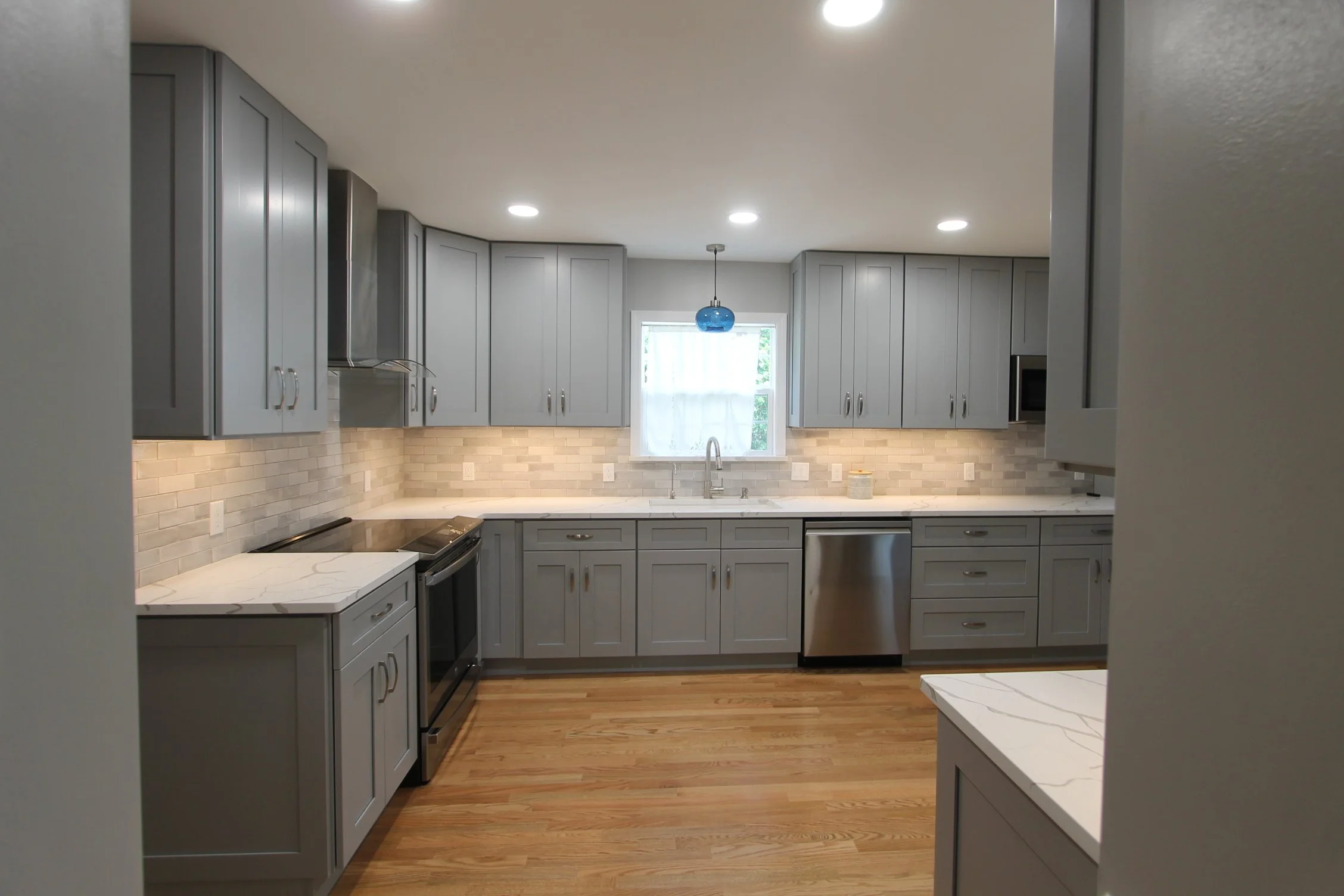 Modern kitchen with gray cabinets, white marble countertops, light wood flooring, and a window with sheer curtains. Recessed ceiling lights and a blue pendant light are visible.