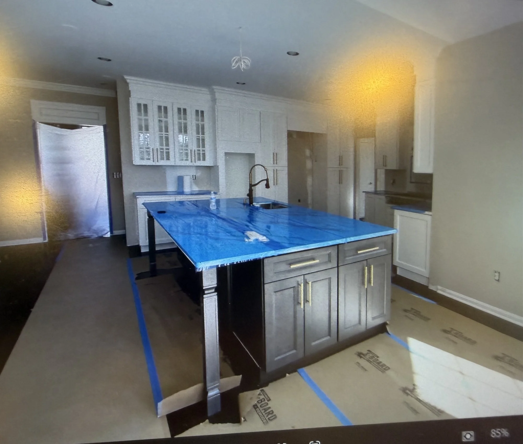 A kitchen with white cabinets, a blue counter, a black faucet, and an island. The room appears to be under construction or renovation with blue painter's tape on the floor and island. There is brown flooring and a door is partially visible on the left side.