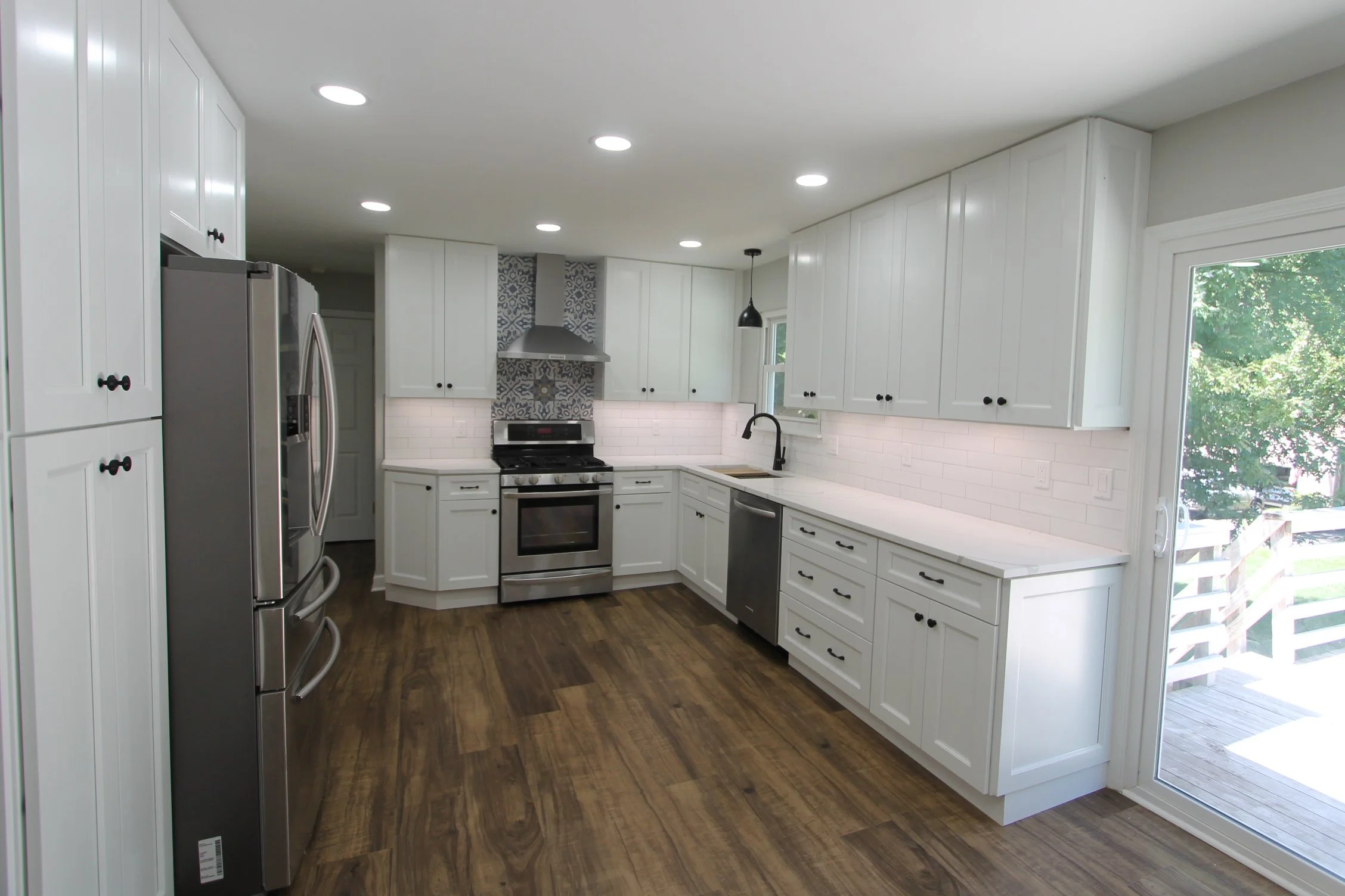 Modern white kitchen with dark wood flooring, stainless steel appliances, white cabinets, a window, and a door leading outside with green trees visible.