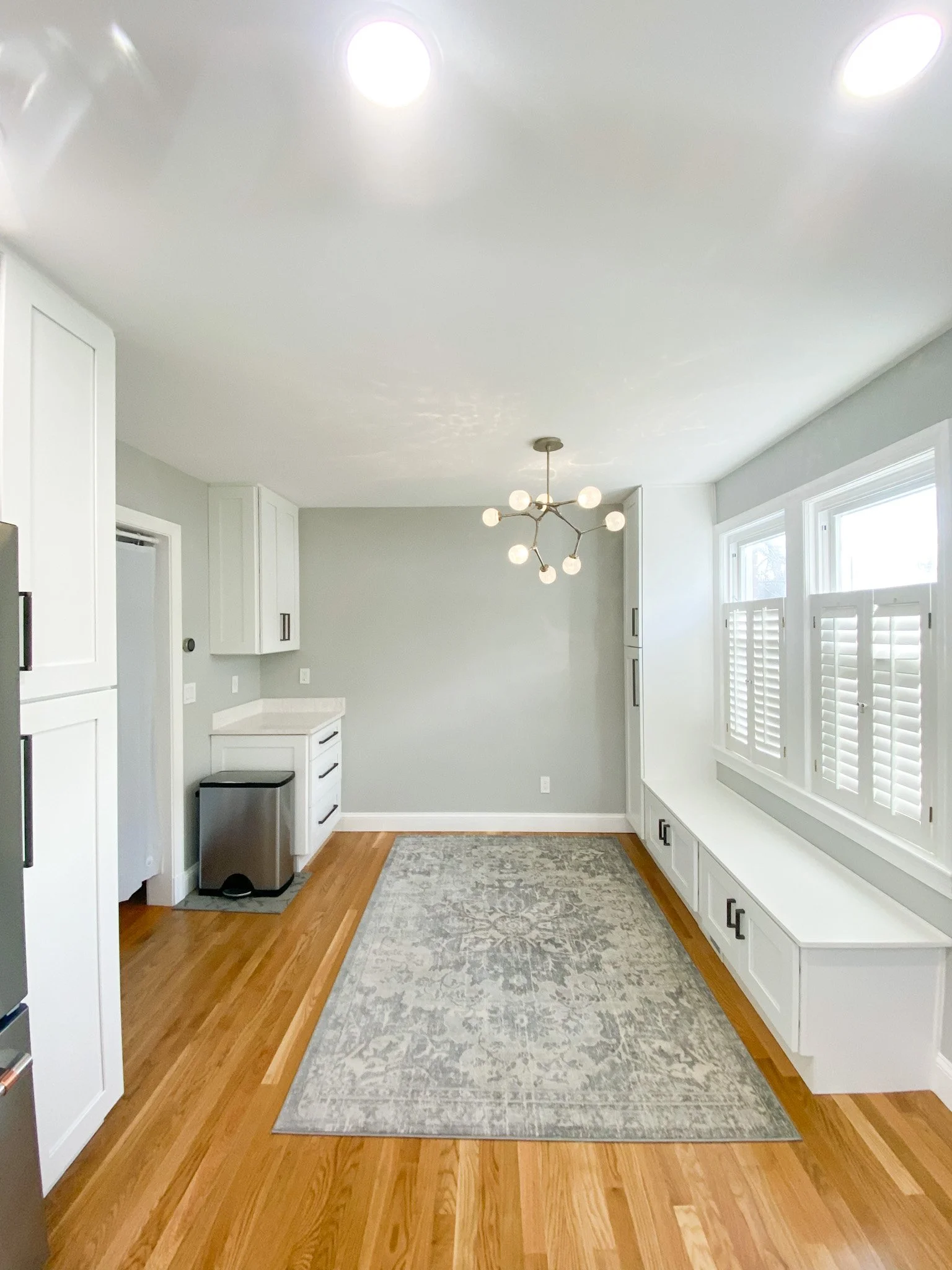 A bright kitchen with white cabinets, large windows with shutters, hardwood floors, a modern chandelier, and a gray patterned rug.