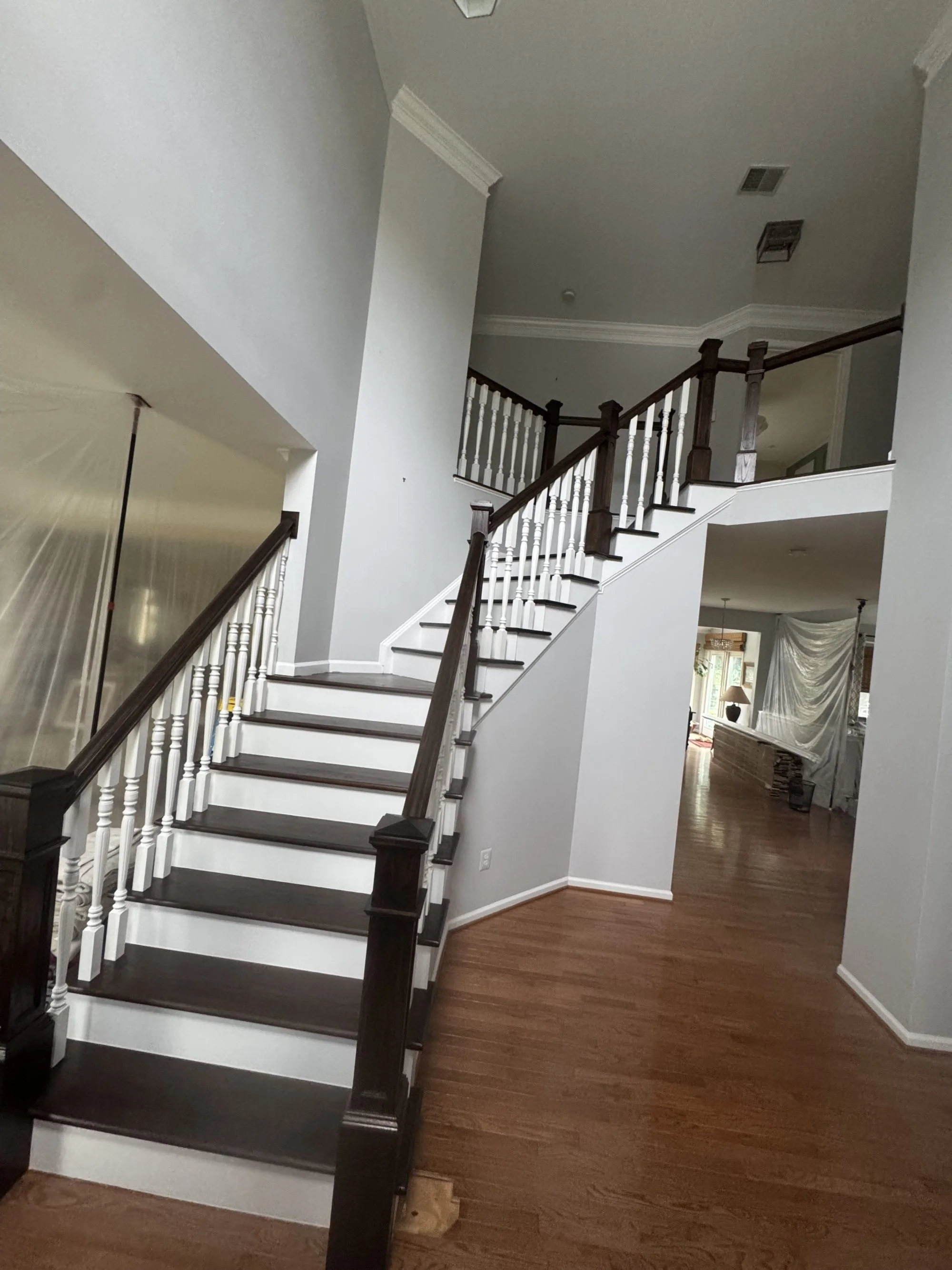 Interior view of a house showing a staircase with dark wooden steps and a white railing with dark wooden handrail, leading to an upper floor. The walls are painted light gray, and the flooring is dark wood. In the background, there's a glimpse of a living room with a window, drapes, and furniture.