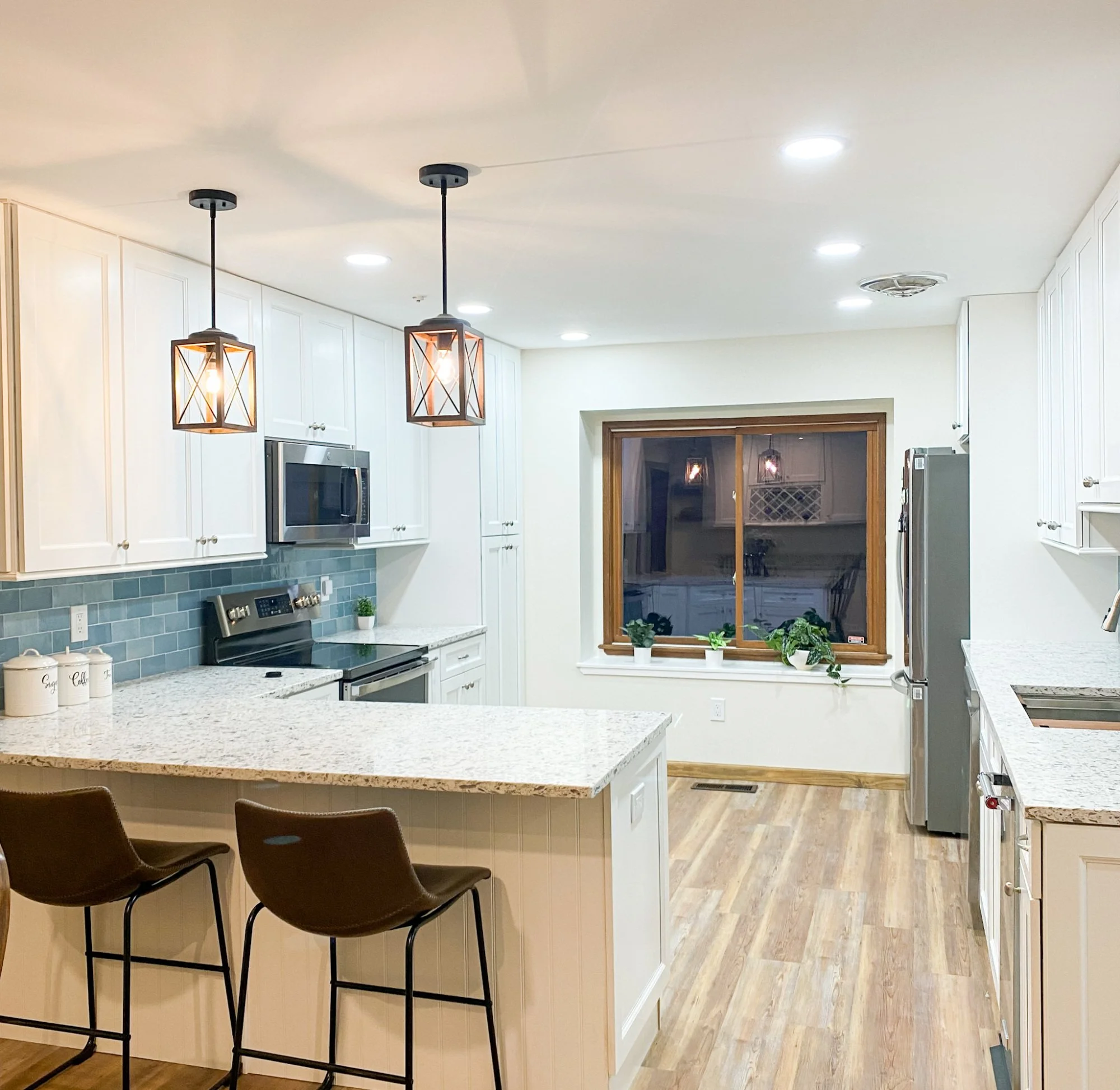Modern kitchen with white cabinets, granite countertops, pendant lights, stainless steel appliances, large window with plants, and wood flooring.