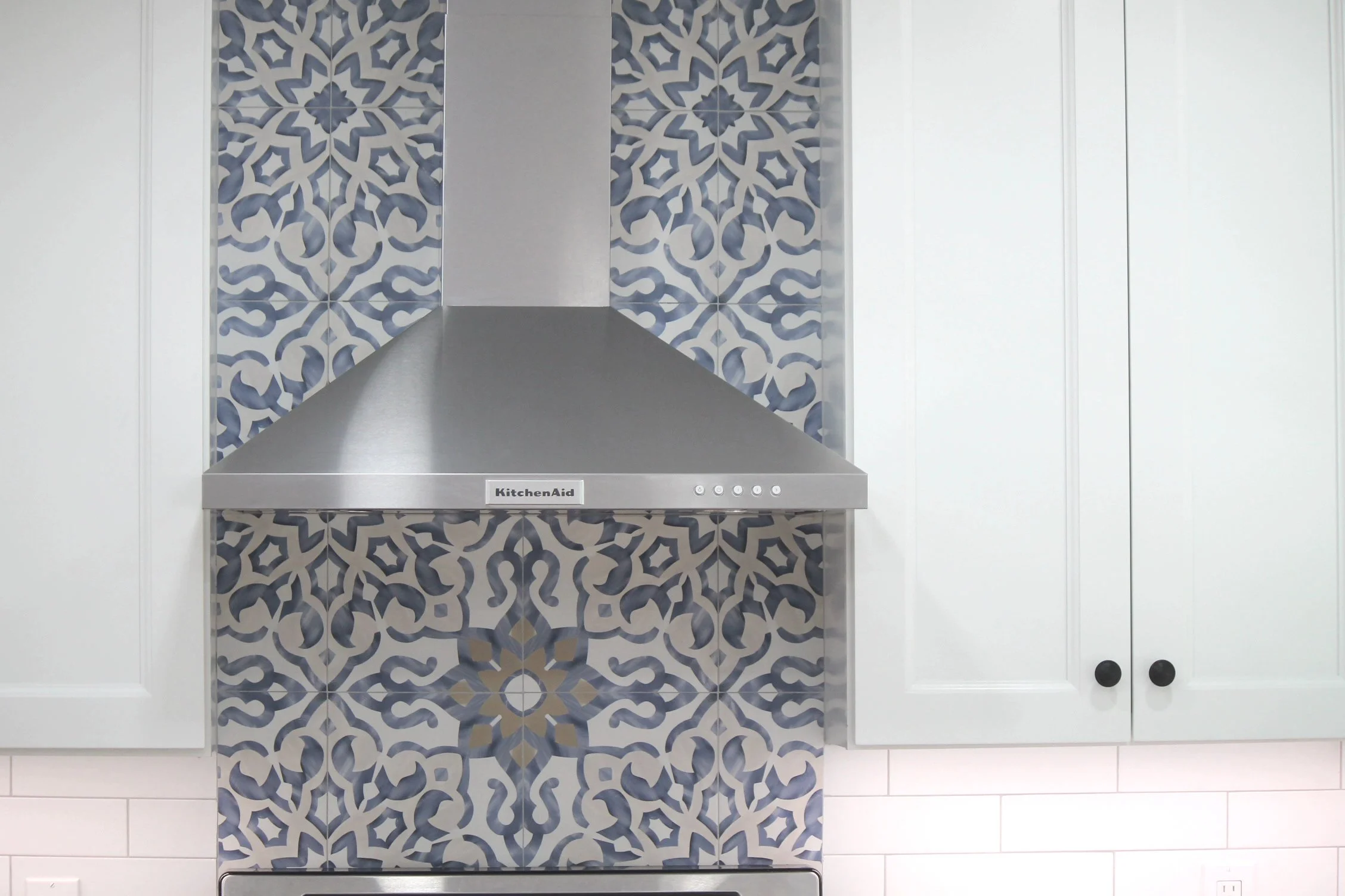Modern kitchen with a decorative backsplash featuring blue and white patterned tiles, stainless steel range hood, white cabinetry with black knobs, and white walls.