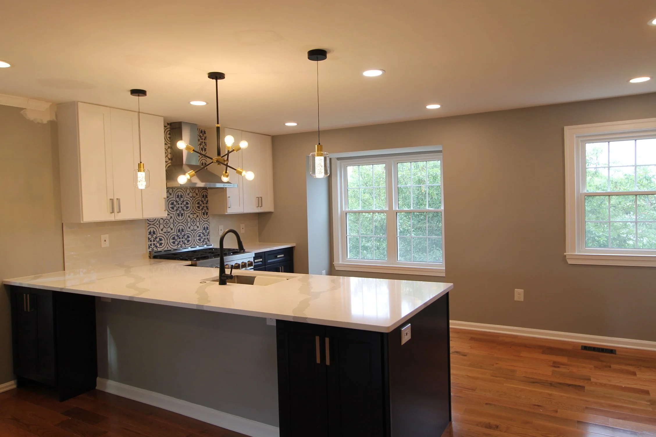 Modern kitchen with white cabinets, a black kitchen island with a white countertop, pendant lights hanging above, a window showing greenery outside, stainless steel range hood, and hardwood floors.