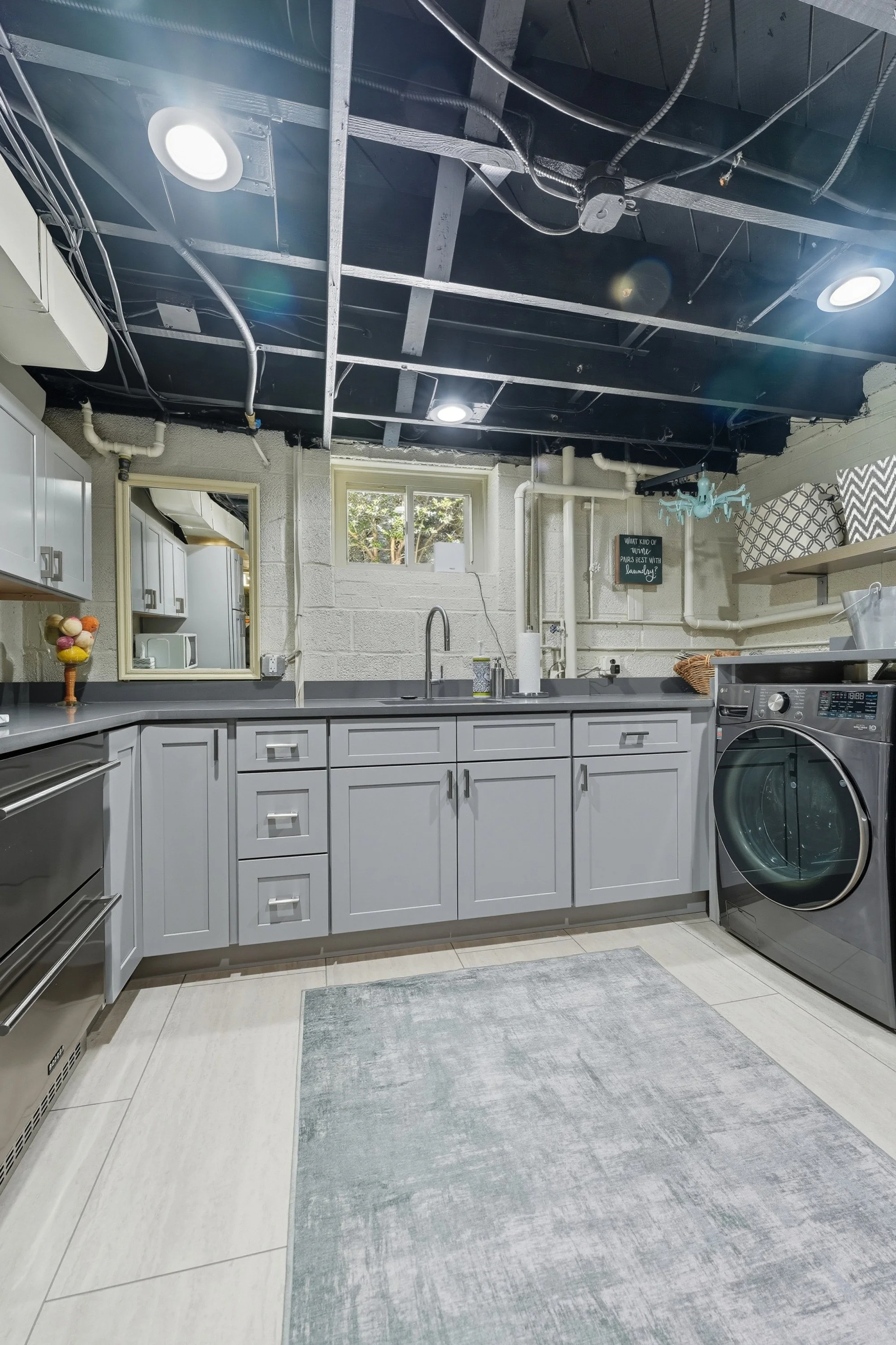 A laundry room with white cabinets, gray countertops, and a washing machine. The room has a small window, a mirror, and a gray rug on the tiled floor. The ceiling is unfinished with visible pipes, wires, and circular recessed lights.