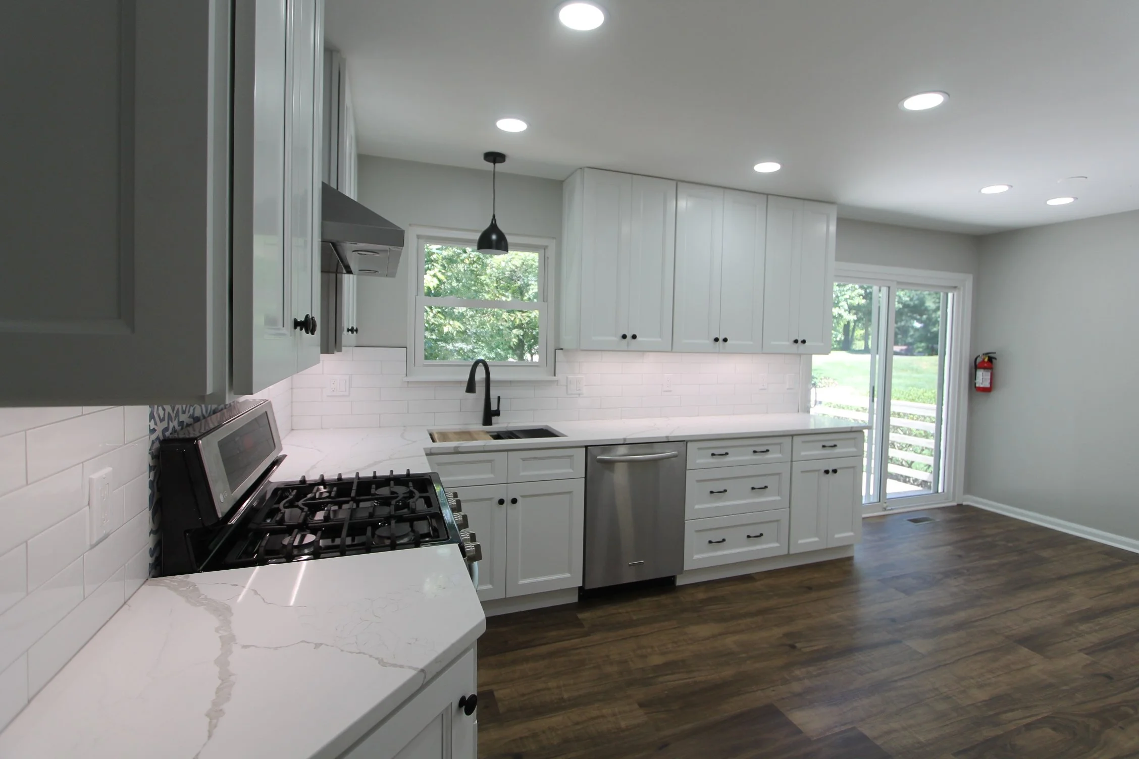 Modern kitchen with white cabinets, a marble countertop, a black pendant light, windows showing greenery outside, a gas stove, and a sliding glass door leading outside.