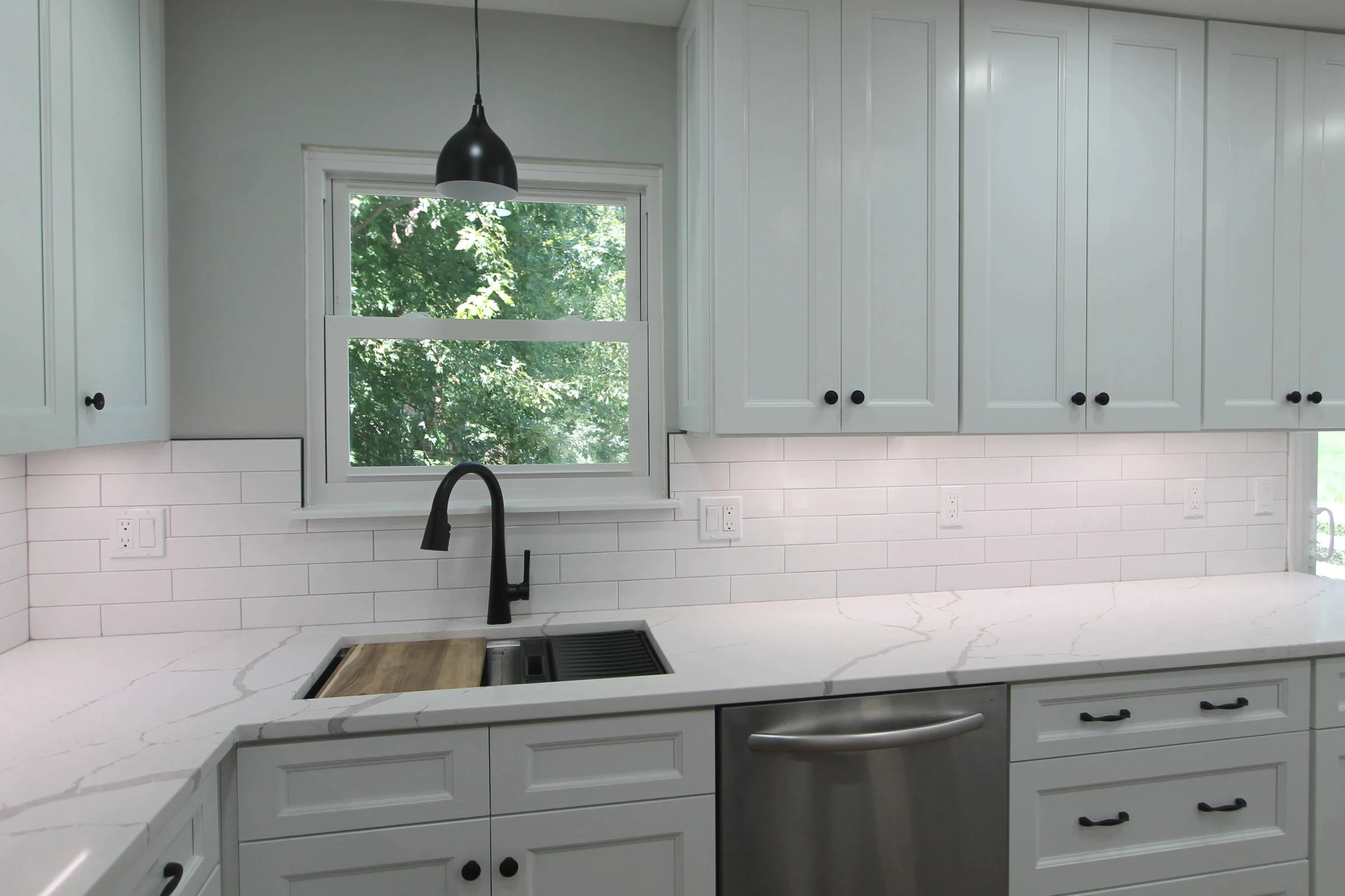 Modern kitchen with white cabinets, marble countertops, black hardware, a black sink faucet, a window with greenery outside, and a black hanging light fixture.